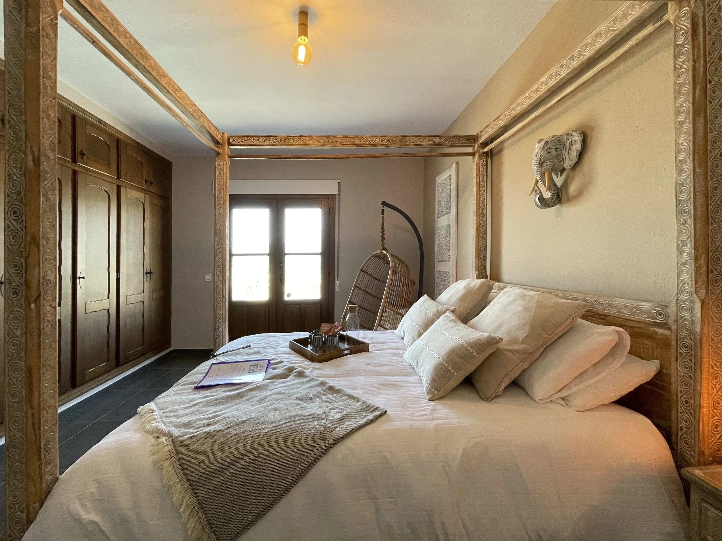 Een slaapkamer met een groot bed dat bedekt is met witte lakens en beige kussens. Aan het hoofdeinde hangt een decoratief masker aan de muur en er is een hangstoel naast het bed. Op de nachttafel ligt een tray met drinken en een boek. De kamer heeft houten details en dubbele deuren die op een balkon uitkomen.
