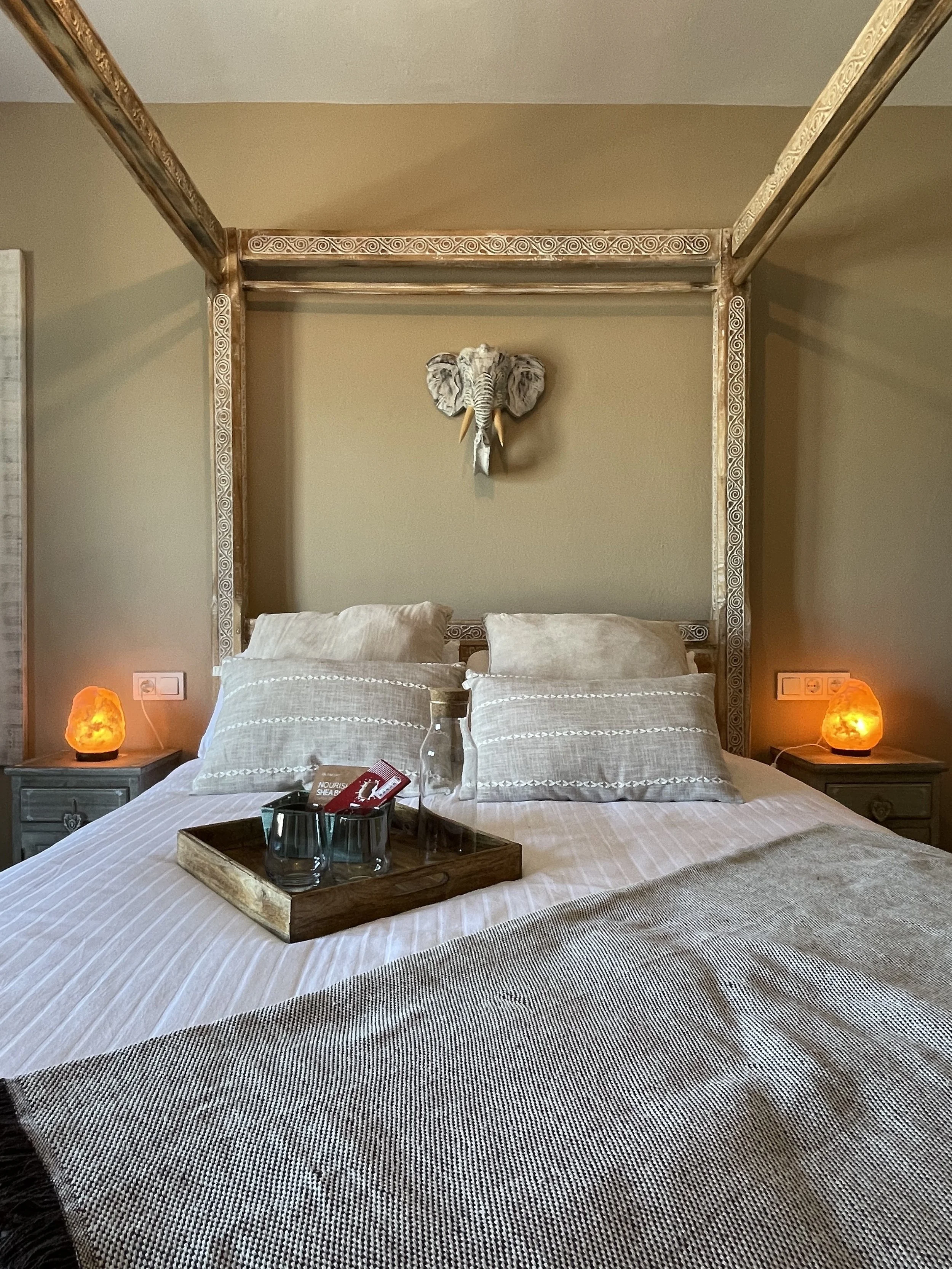Een slaapkamer met een groot bed, beige beddengoed en een houten tray met glazen, flessen en kleine artikelen. Twee nachtkastjes met oranje zoutlampen, een muurdecoratie van een olifant hoornkop, en een hoek met stopcontacten.