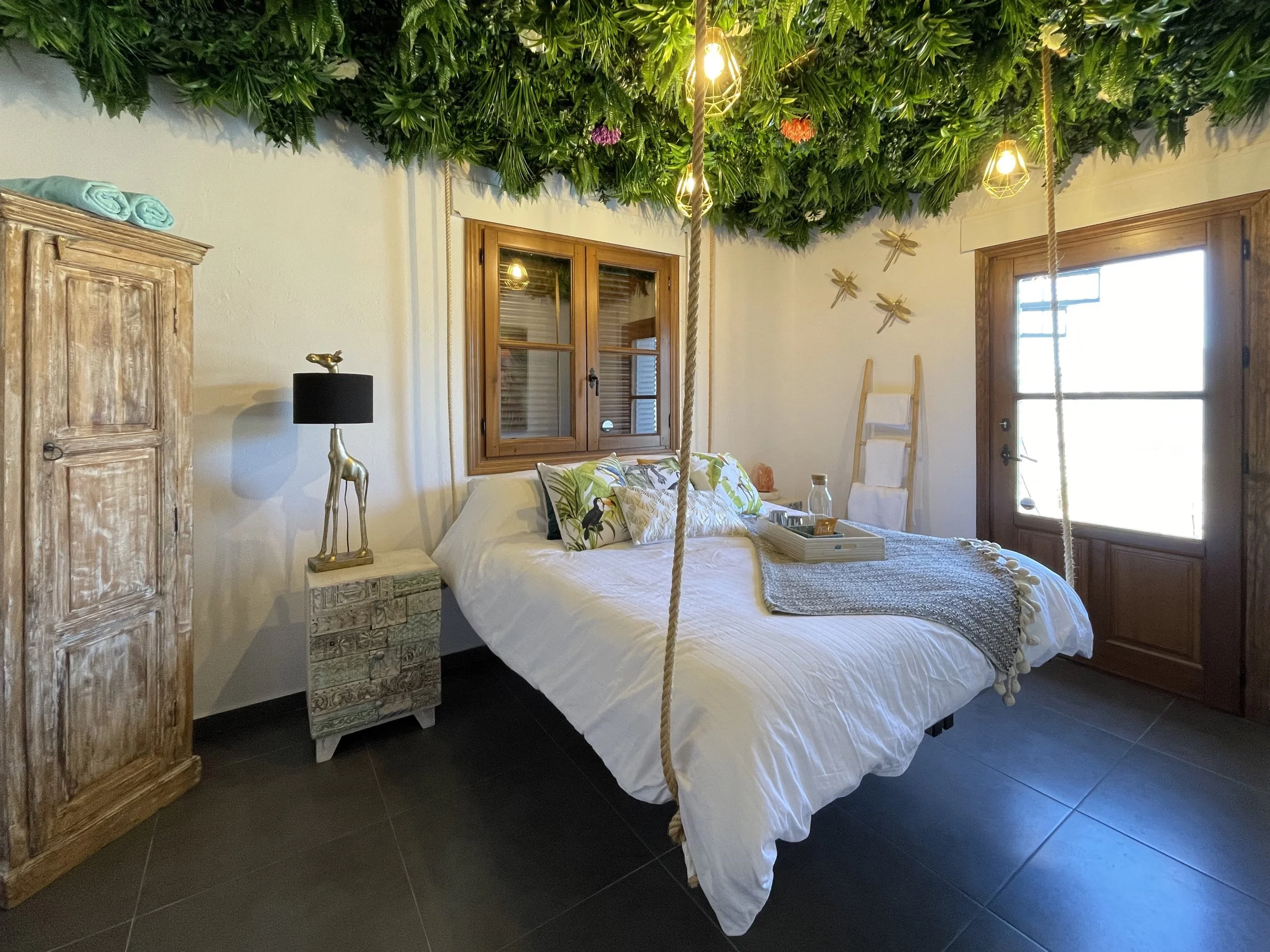 Een slaapkamer met een bed, een houten kast, een nachtkastje met een lamp, een raam en een deur. Het plafond is versierd met groene planten en hangende lampen.