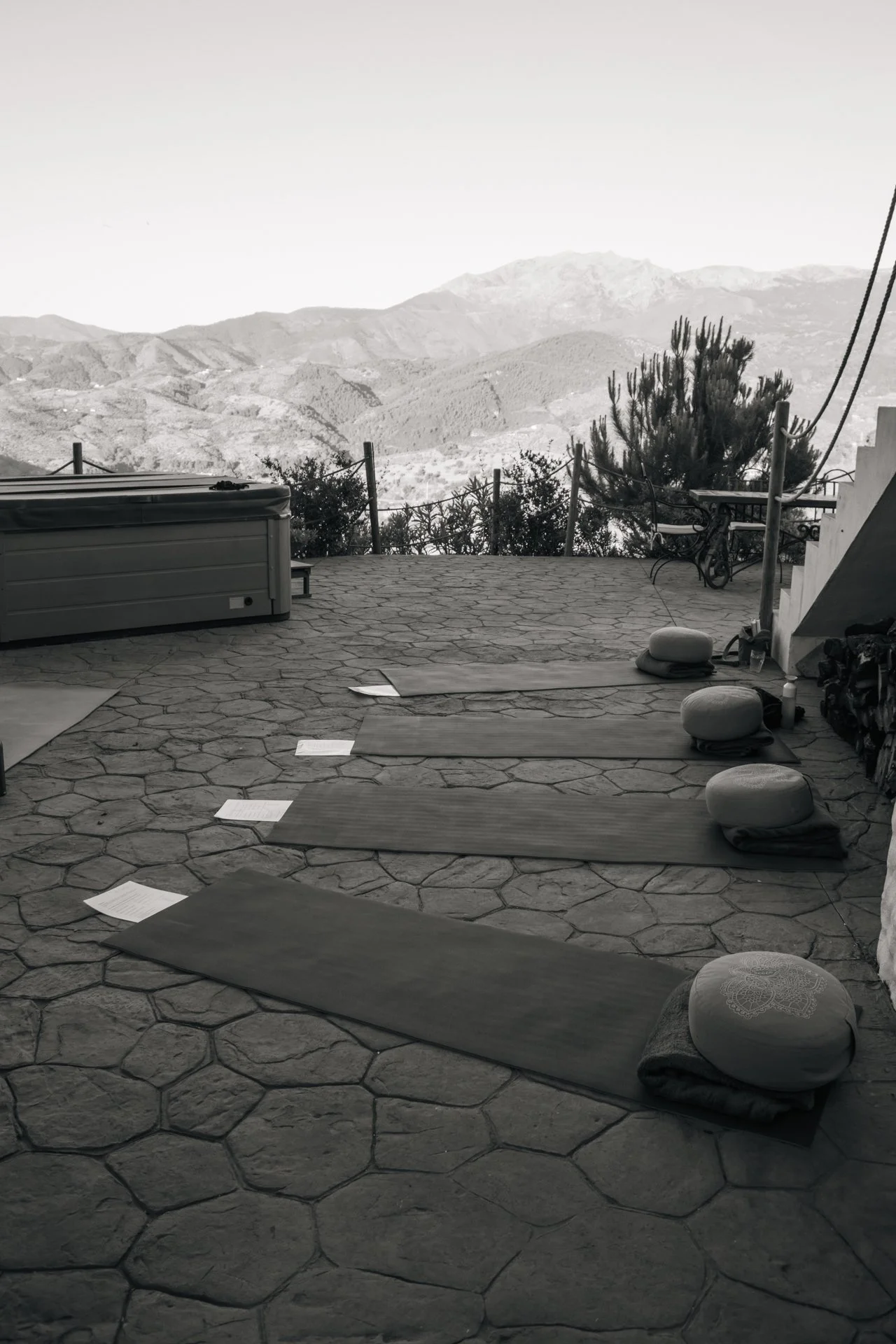 Binnenruimte met yogamatten, meditatiekussens en papieren op de stenen vloer, uitzicht op bergen in de verte, mogelijk een meditatie- of yogaruimte.