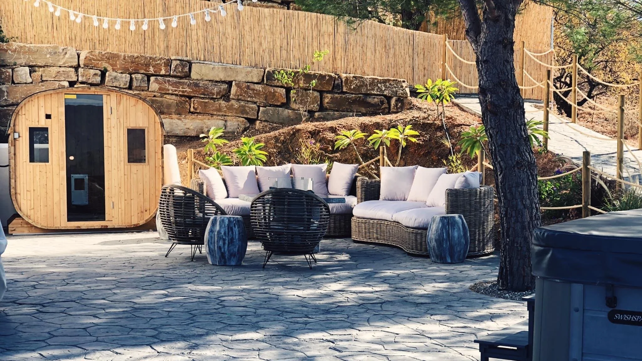 Een zithoek met een loungebank en bijzettafels, omgeven door bomen en een tuinmuur, in een buitenomgeving.