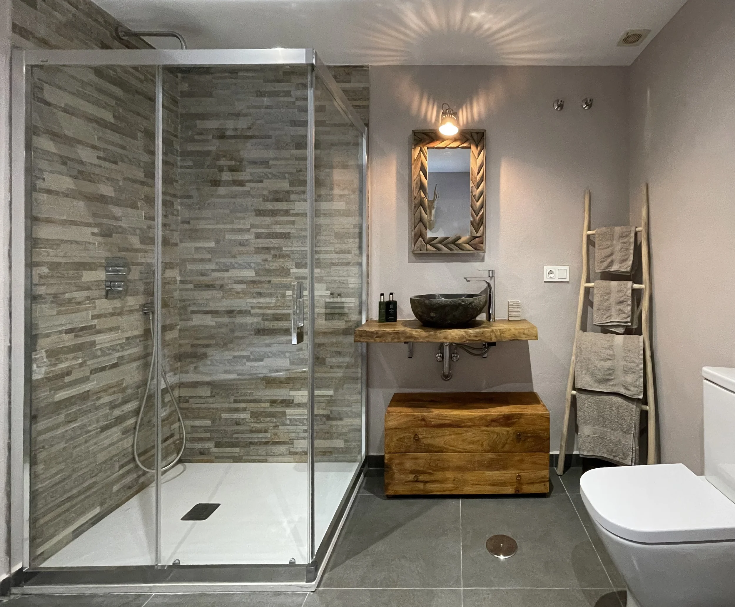 Moderne badkamer met inloopdouche met stenen muurstuc, houten kast met waskommen, spiegel, handdoekenrek en douchegel.