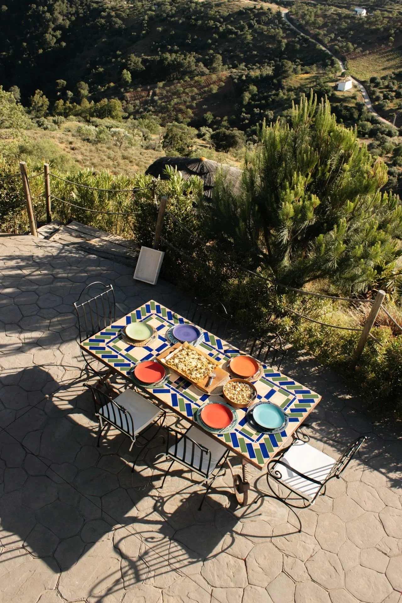 Tuin met een gedekte tafel met kleurrijke borden, omringd door groene planten en een landschap met heuvels en olijfbomen in de achtergrond.