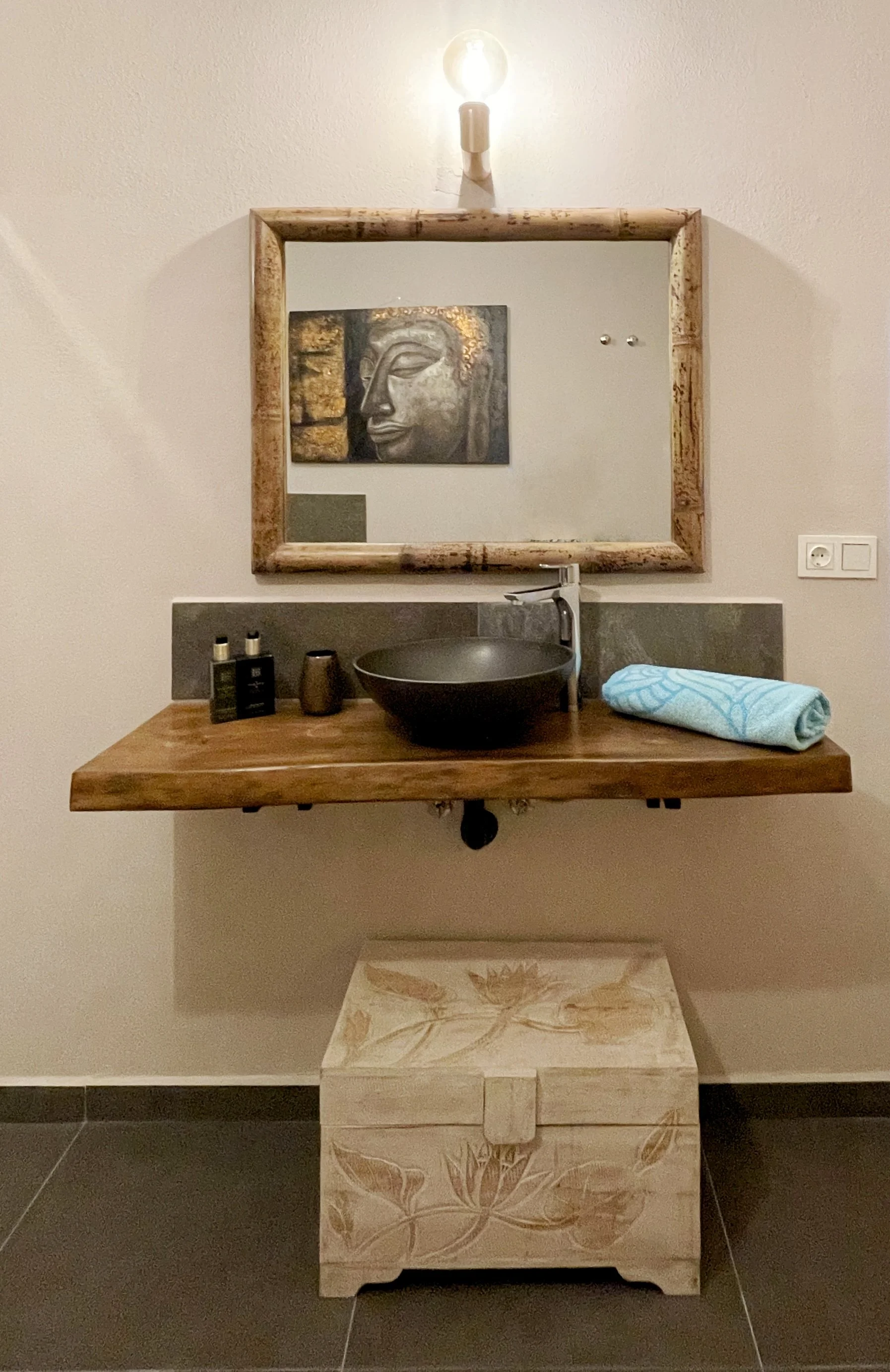 Een badkamer met een houten wastafel, een ronde zwarte waskom, een spiegel met houten rand, een kunstwerk van een vredessymbolische afbeelding, een lichtarmatuur boven de spiegel, handdoek, en verzorgingsproducten op de rand.
