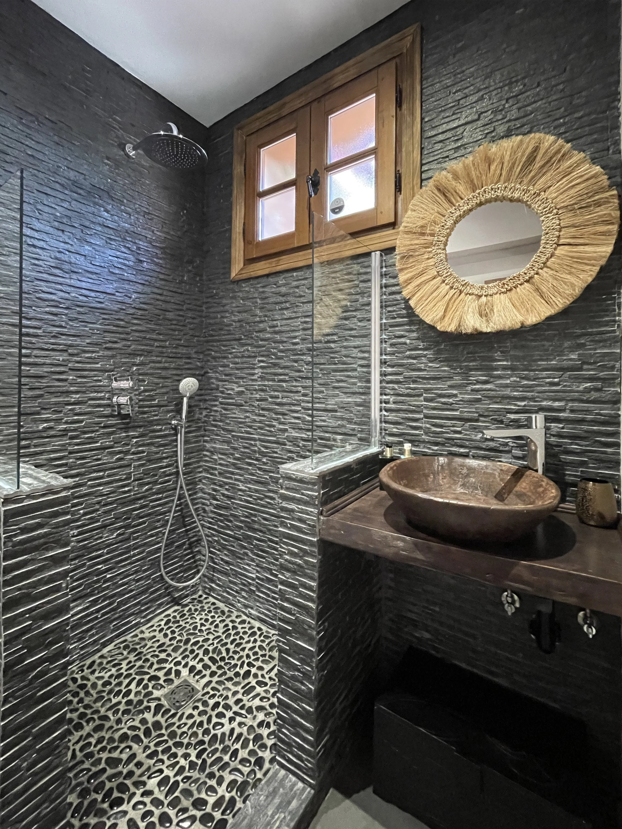 Moderne badkamer met zwarte steenachtige muurtegels, een inloopdouche met een regen douchekop, een ronde aardewerken wastafel op een houten onderstel, een grote ronde spiegel met een omhulsel van touw, en een klein raam met houten kozijn.
