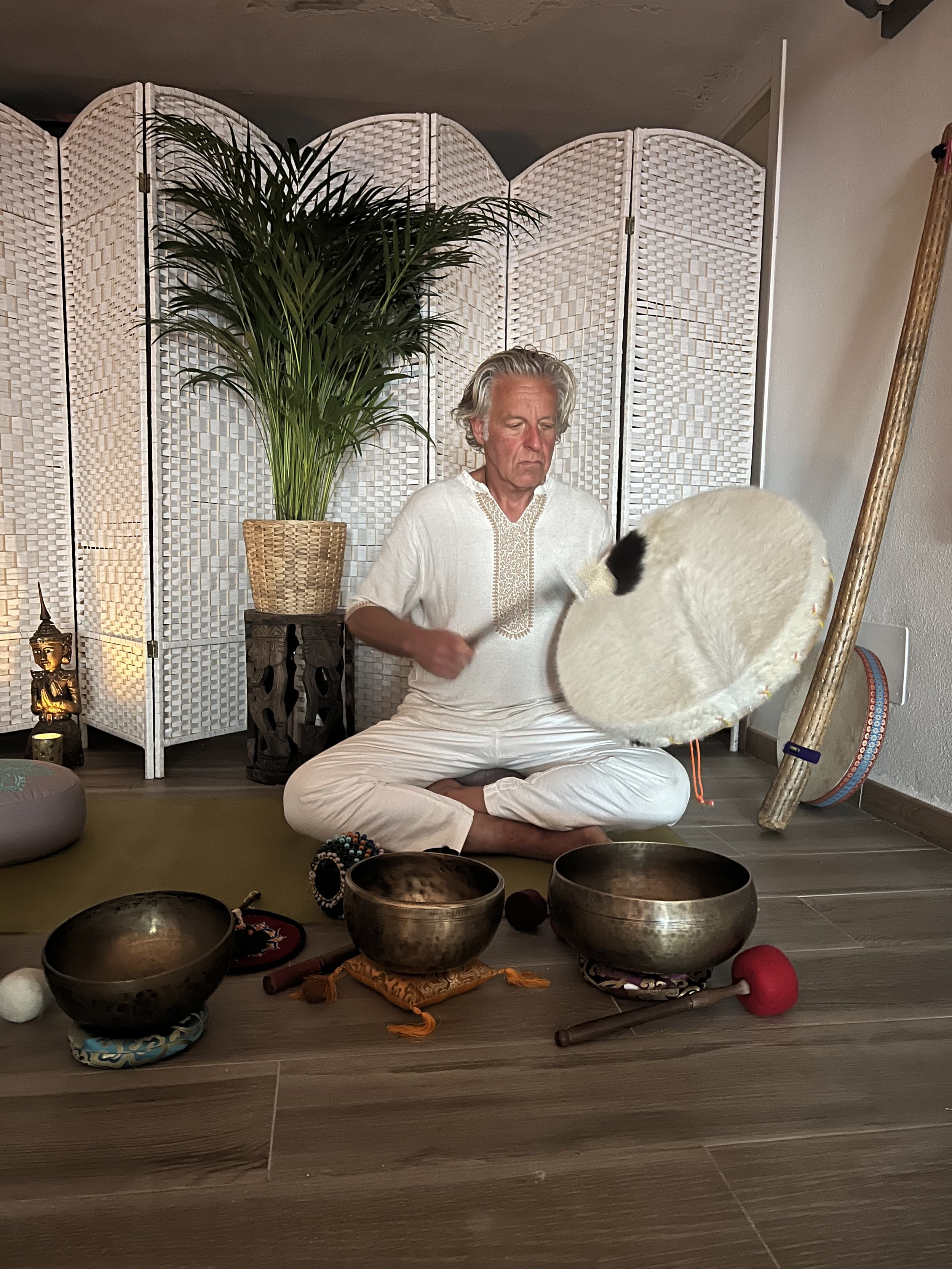 Een man zit geknield op de vloer, omringd door Tibetaanse klankschalen, terwijl hij zich voorbereidt op een klankmeditatie. De achtergrond bevat een witte kamerscherm en een grote groene plant.