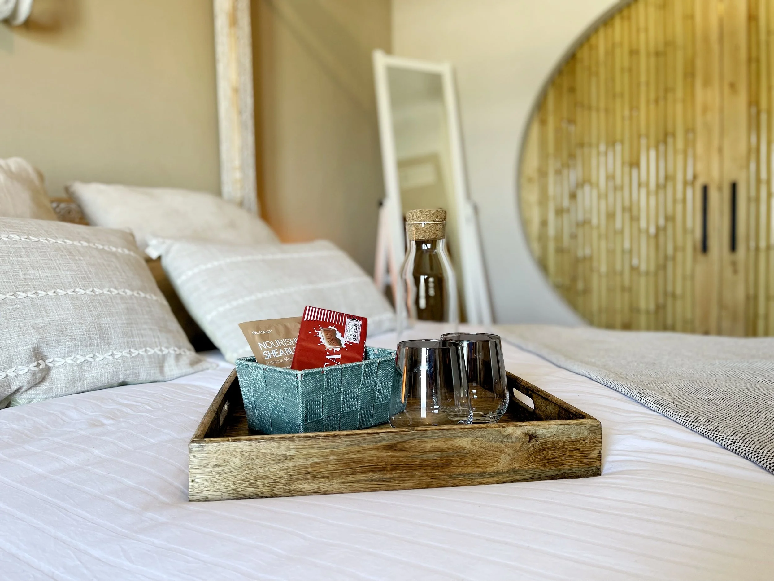 Een hotelkamer met een bed, kussen en een houten dienblad op het bed met schaaltjes en glazen, een spiegel en een wand met houten decoratie.