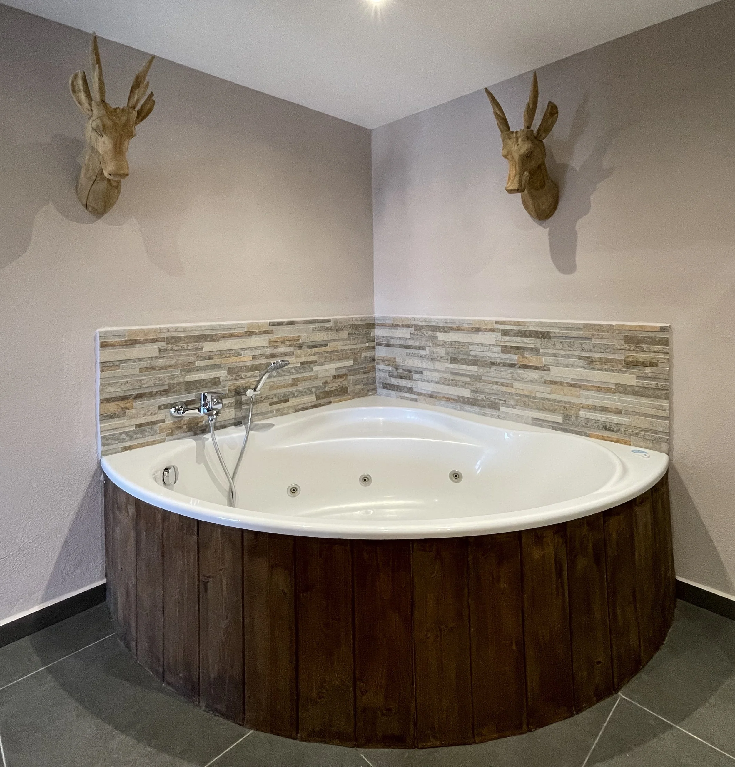 Een badkamer met een hoekbad, houten wastafelmeubel en een betegelde muur onderaan.