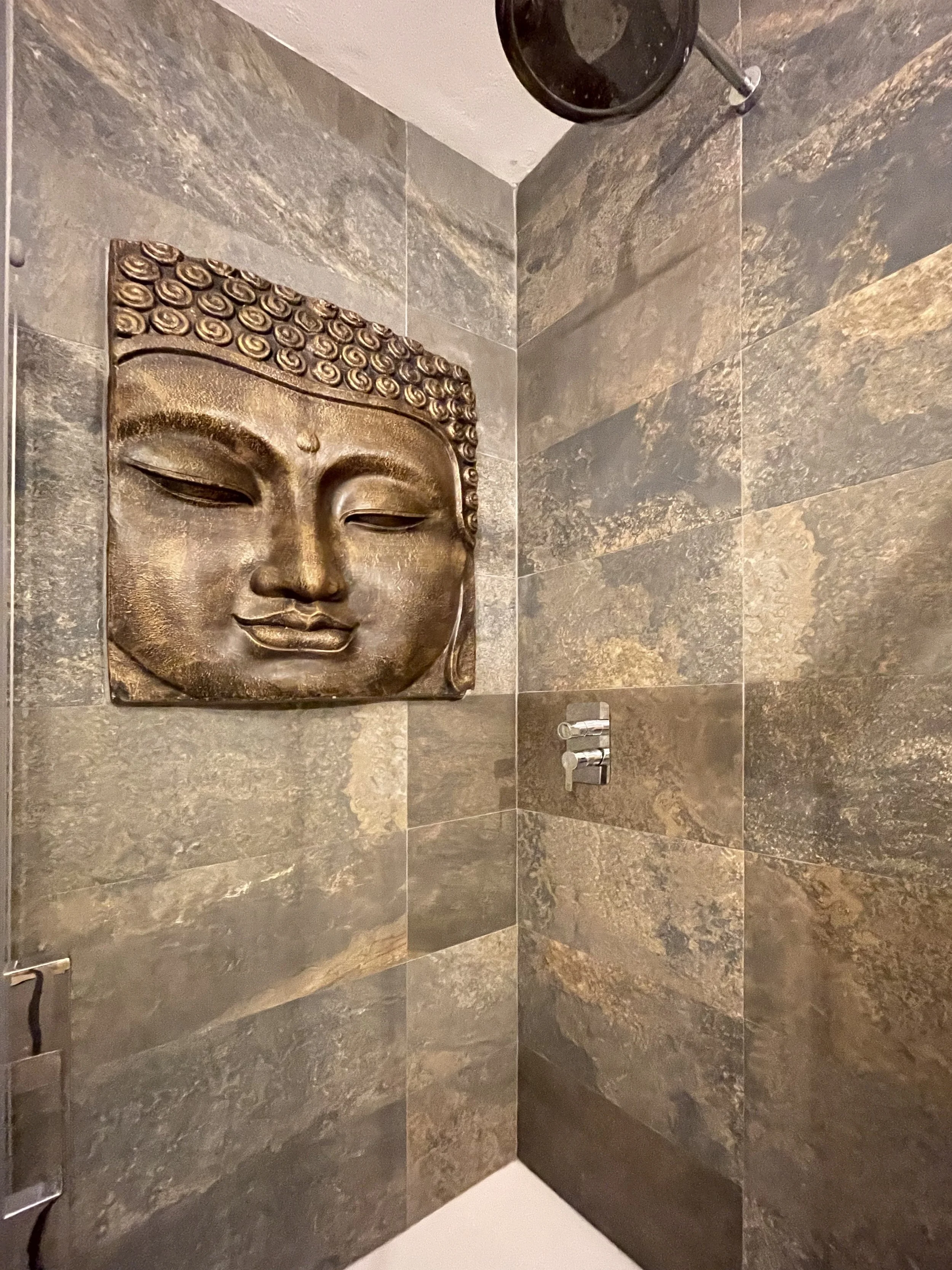 Een gouden boeddhabeeld met gesloten ogen op een muur met stenen tegels in een badkamer. En er ligt een zwarte emmer aan het plafond.