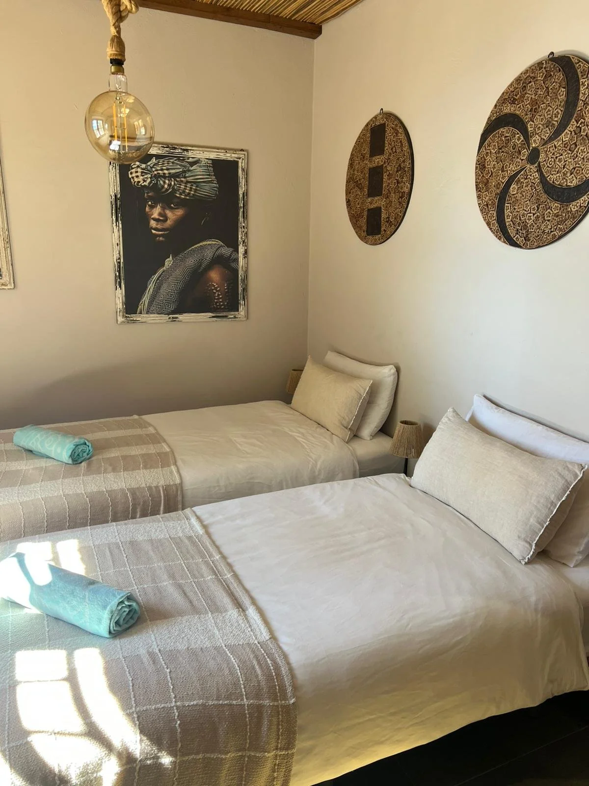 Twee geprepareerde bedden met witte lakens en beige dekens, met op elke bed een kussen en een opgerolde turquoise handdoek. In de hoek van de kamer hangen drie decoratieve wandplaten en een portret van een persoon met een traditioneel hoofddeksel en sieraden. Boven de bedden hangt een glazen hanglamp met een groot gloeilamp.