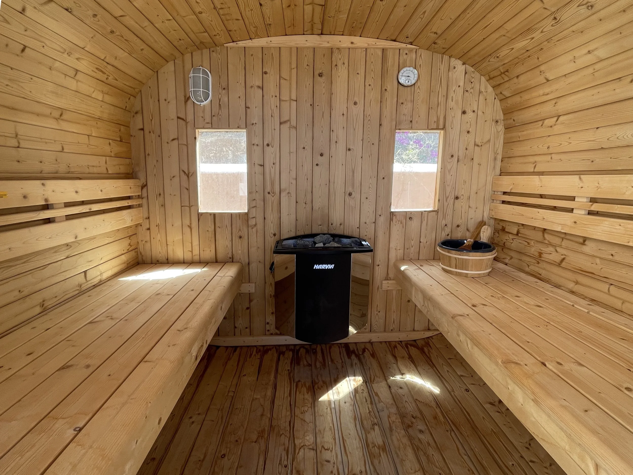 Binnenkant van een houten sauna met rond plafond, twee kleine ramen, een verwarming met stenen, en een houten emmer.