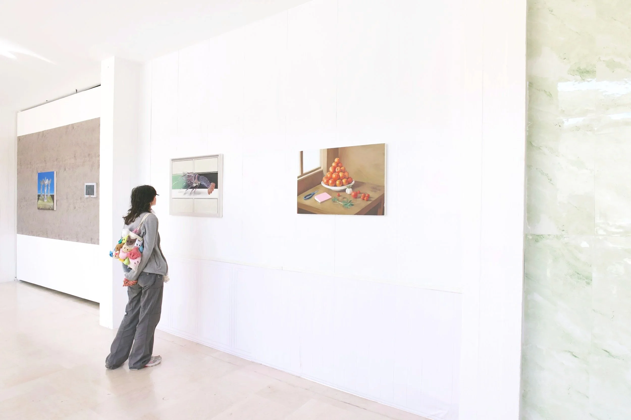 A woman with glasses, grey hoodie, and grey pants carrying plush toys, stands in front of a white gallery wall with three paintings, including a still life of tomatoes, a portrait of a lobster, and a landscape of trees or mountains.