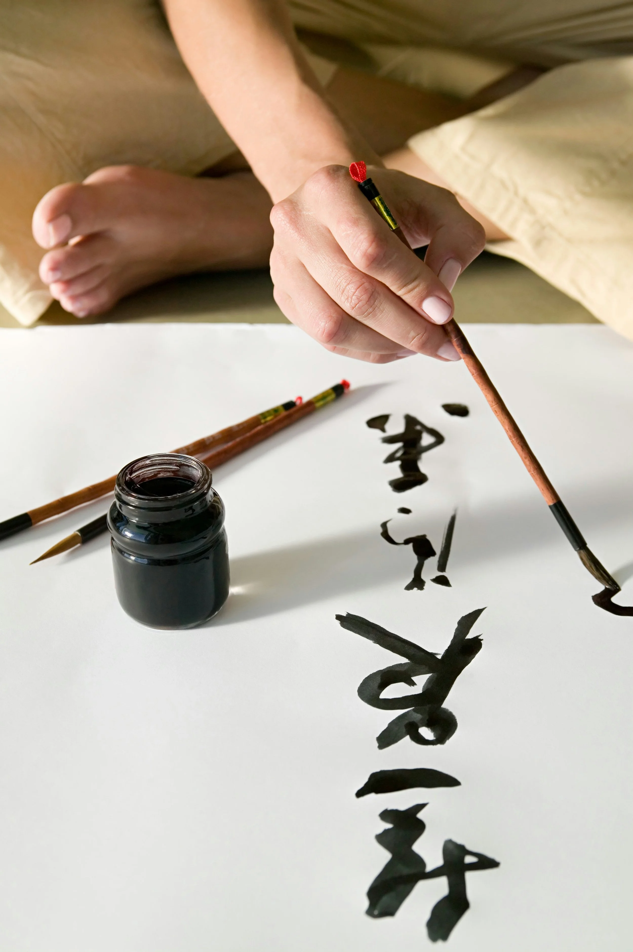 A hand is holding a brush and writing Chinese characters with black ink on paper. An ink bottle and another brush are on the table.