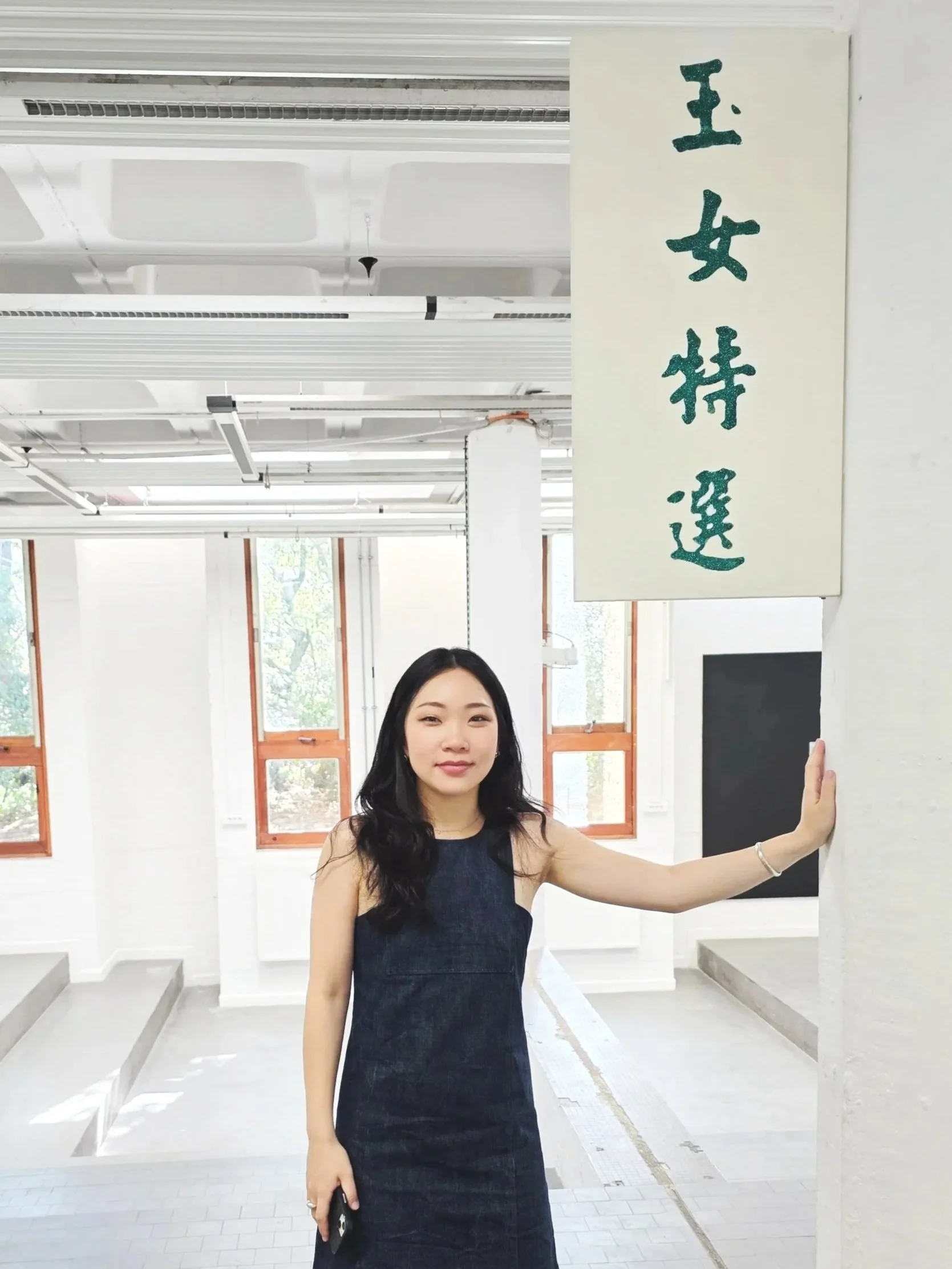 A woman in a black dress standing indoors with her hand on a white pillar and smiling in front of a white wall and windows, with a vertical white sign with green Chinese characters on it.