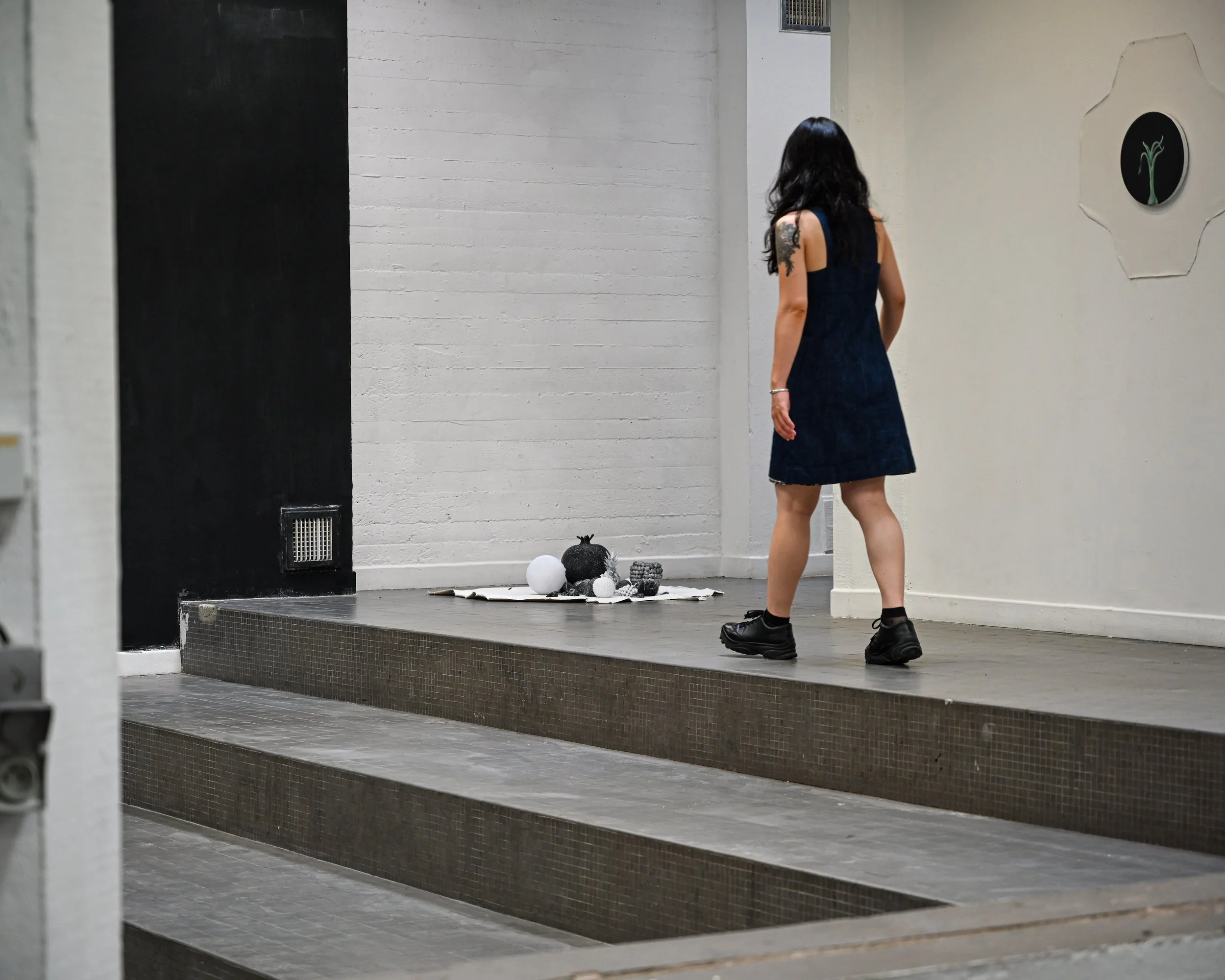 A woman with dark hair wearing a black dress and black shoes is standing on a small stage in an indoor space, with her back turned to the camera. There are decorations on the floor nearby and a black and white circular wall decorations.