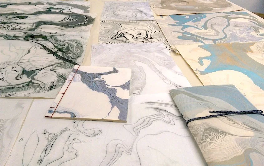 Multiple sheets of marbled paper with various swirling patterns in black, white, gray, and blue tones laid out on a table.