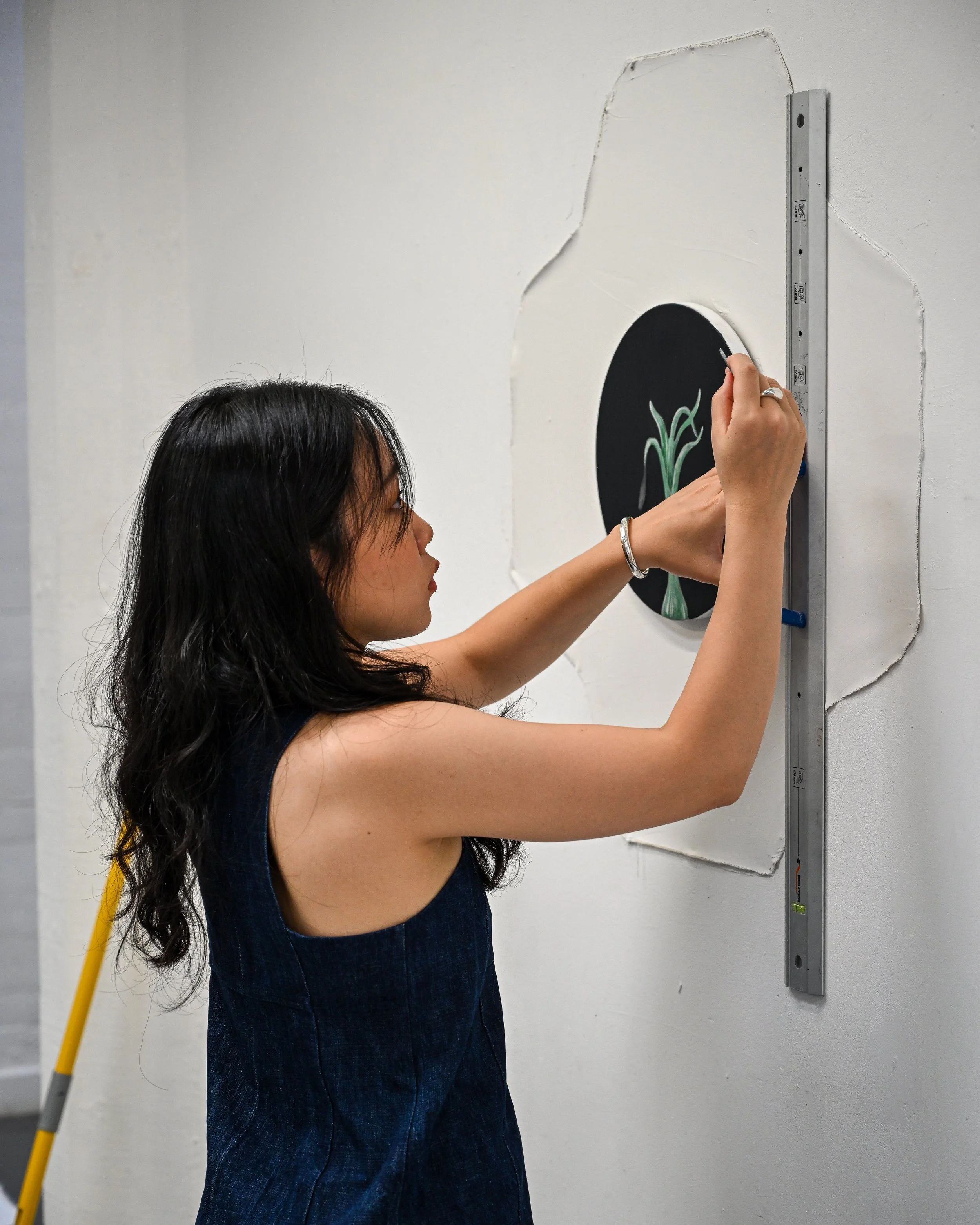 A woman with long dark hair is using a pencil to draw on a circular chalkboard mounted on a white wall. The wall has an uneven cutout around the chalkboard, and there is a yellow ladder in the background.