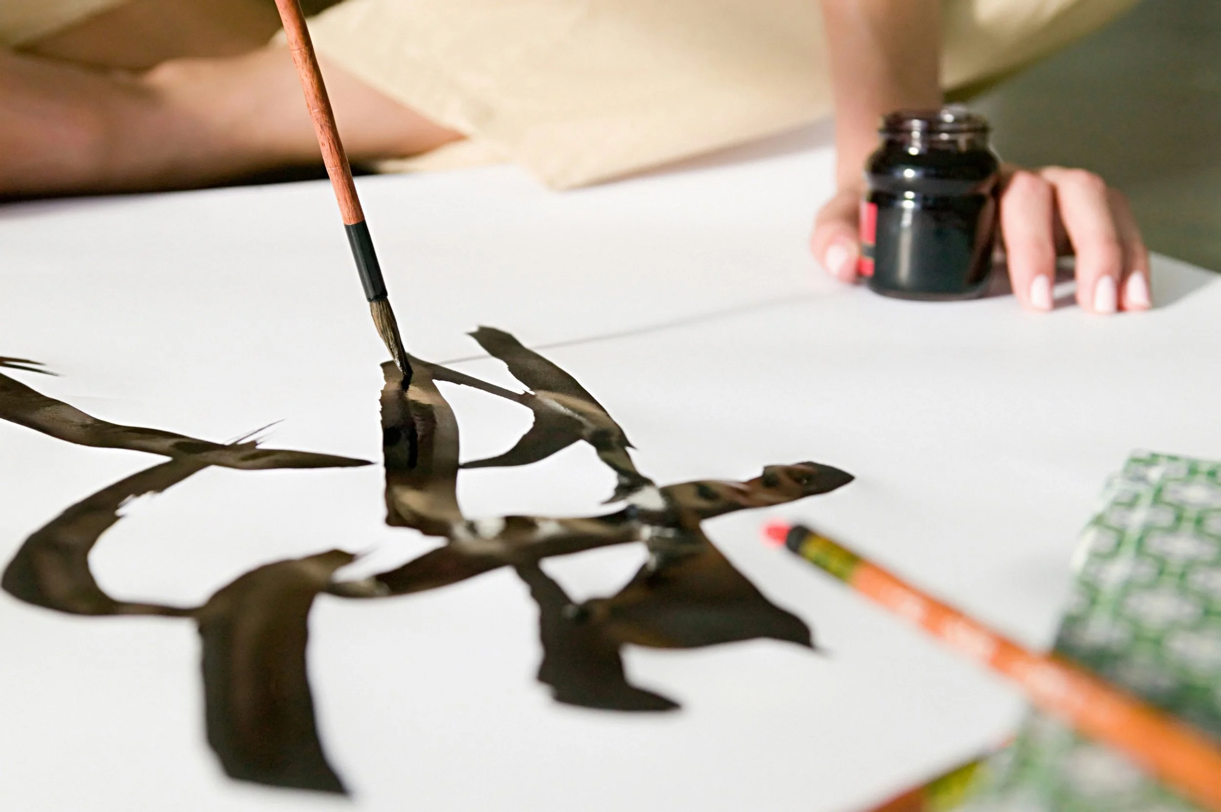 A person using a paintbrush to create a dark ink or paint design on white paper, with a small black ink bottle and some colored pencils nearby.