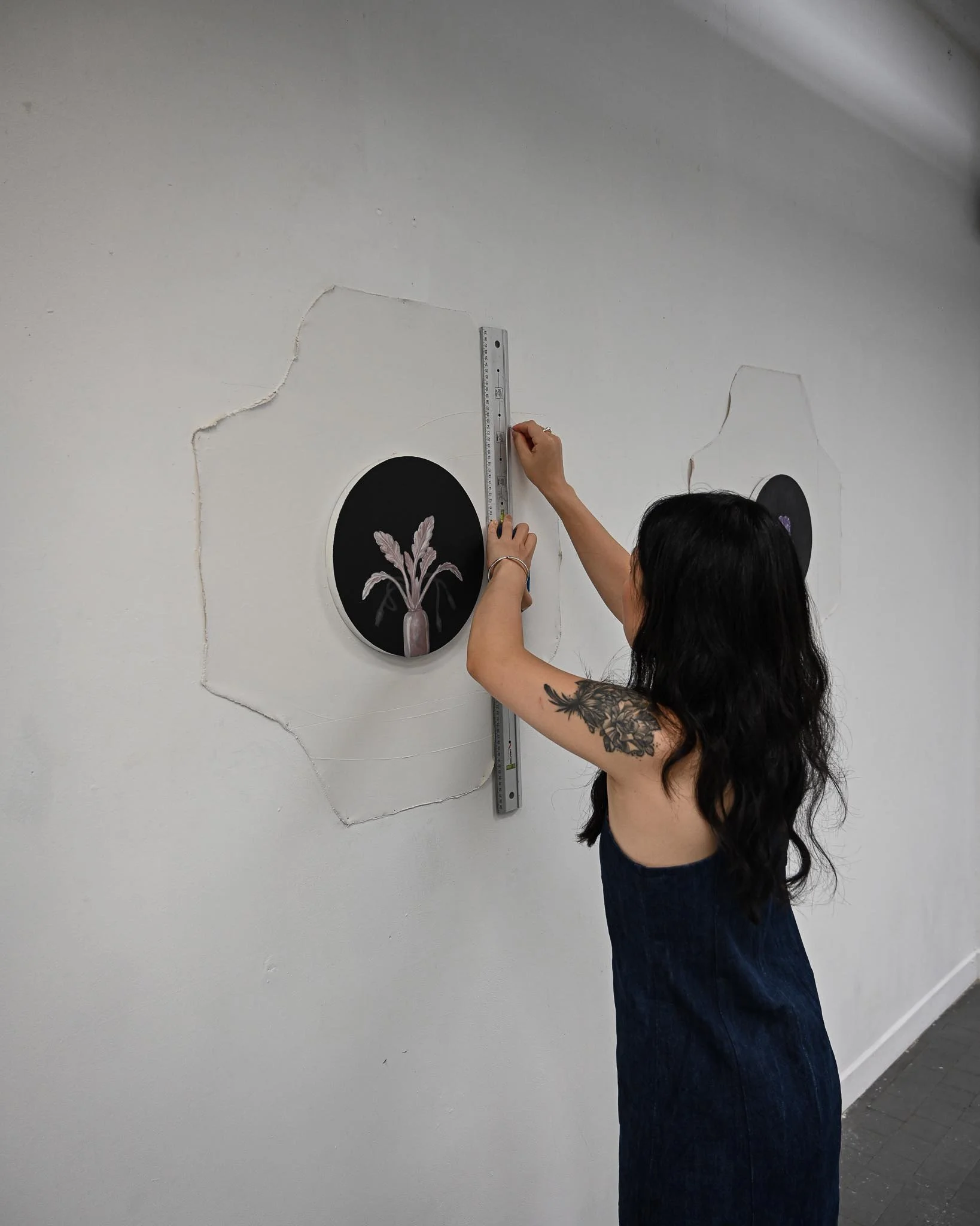 Person measuring a crack on a white wall near a black circular artwork with a white leaf pattern, standing in a room with gray tile flooring.