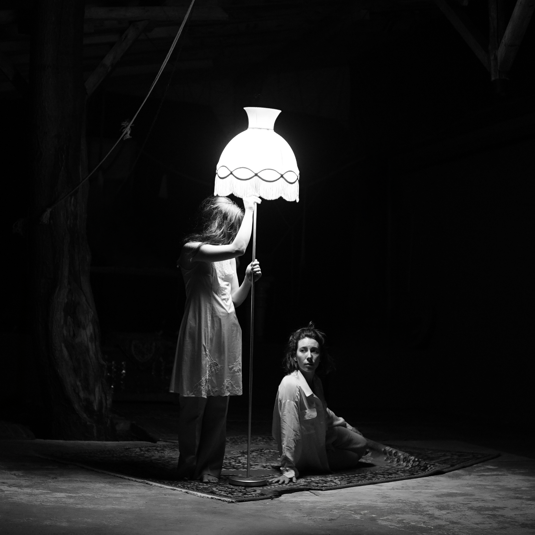 Two women in vintage-style clothing indoor in a dimly lit space, one standing behind a lit lamp and the other sitting on a rug.
