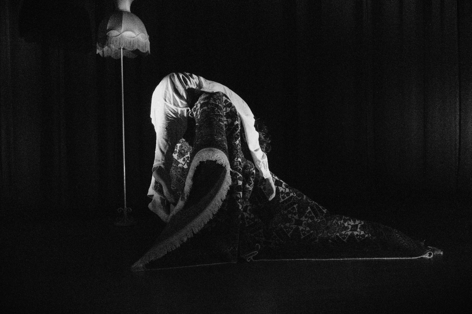 A black and white photograph of two performers on a stage, one person wearing a long patterned dress and the other dressed in light-colored clothing, with a vintage standing lamp with a fringed lampshade in the background.