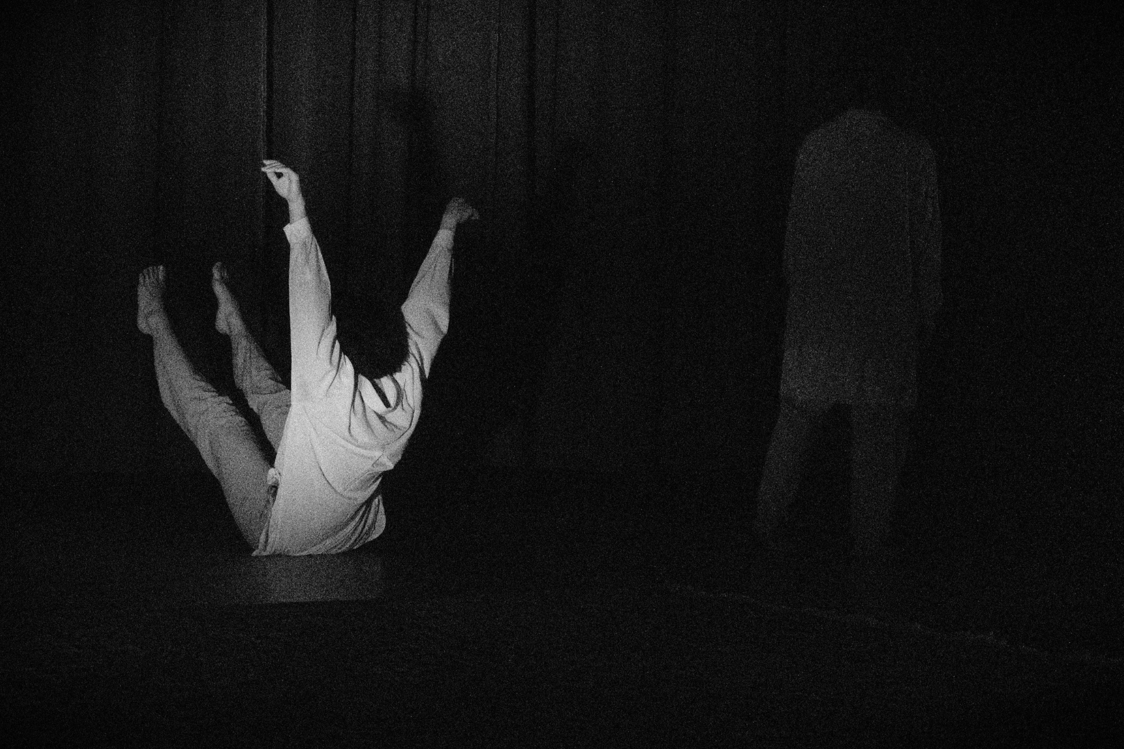A person in white clothing appears to be falling backward or down onto the floor in a dark room, with another person standing nearby, partially visible.