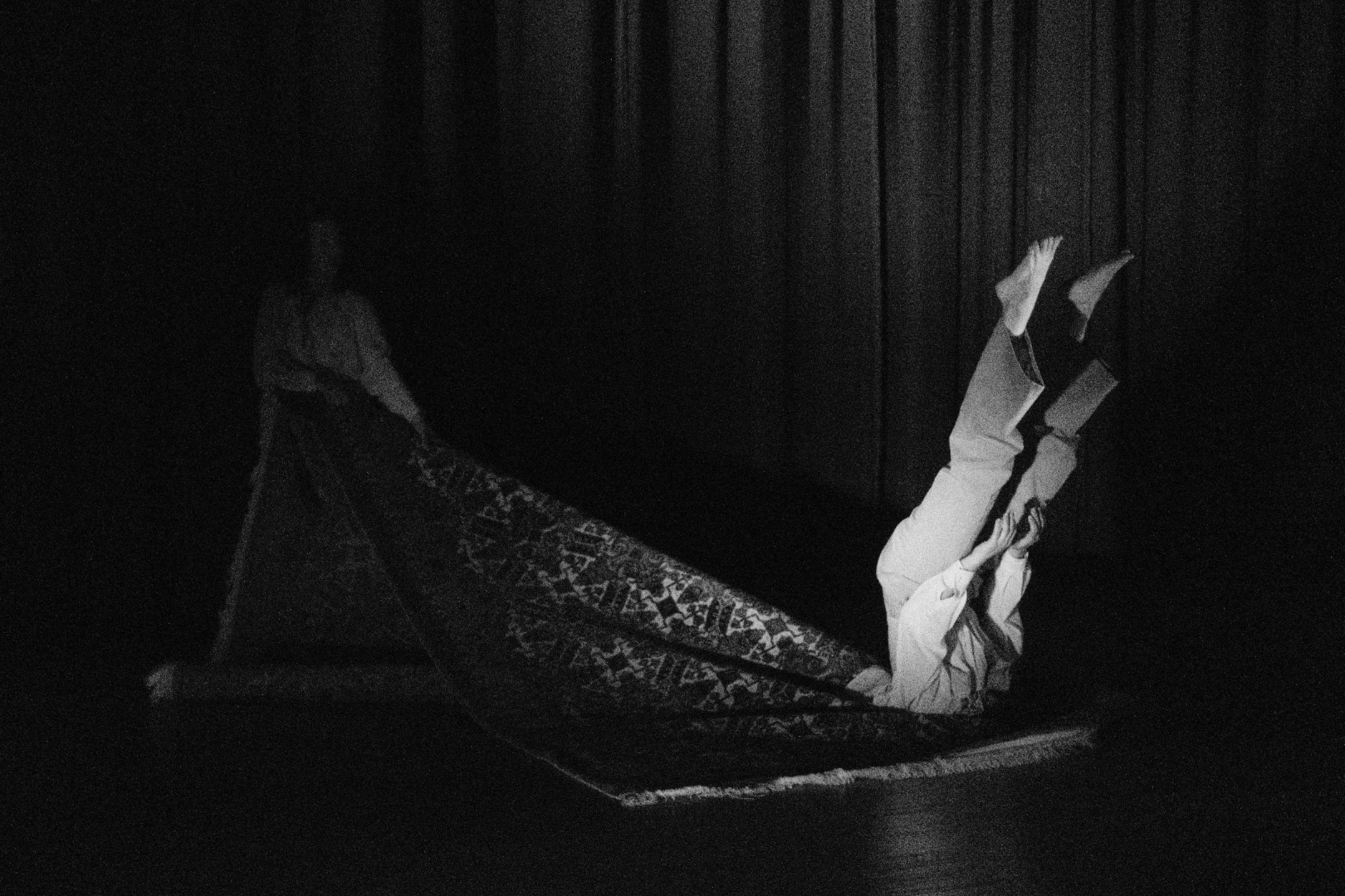 A person lying on a patterned rug with their legs raised against a wall, holding a book, in a dark room with wooden walls.