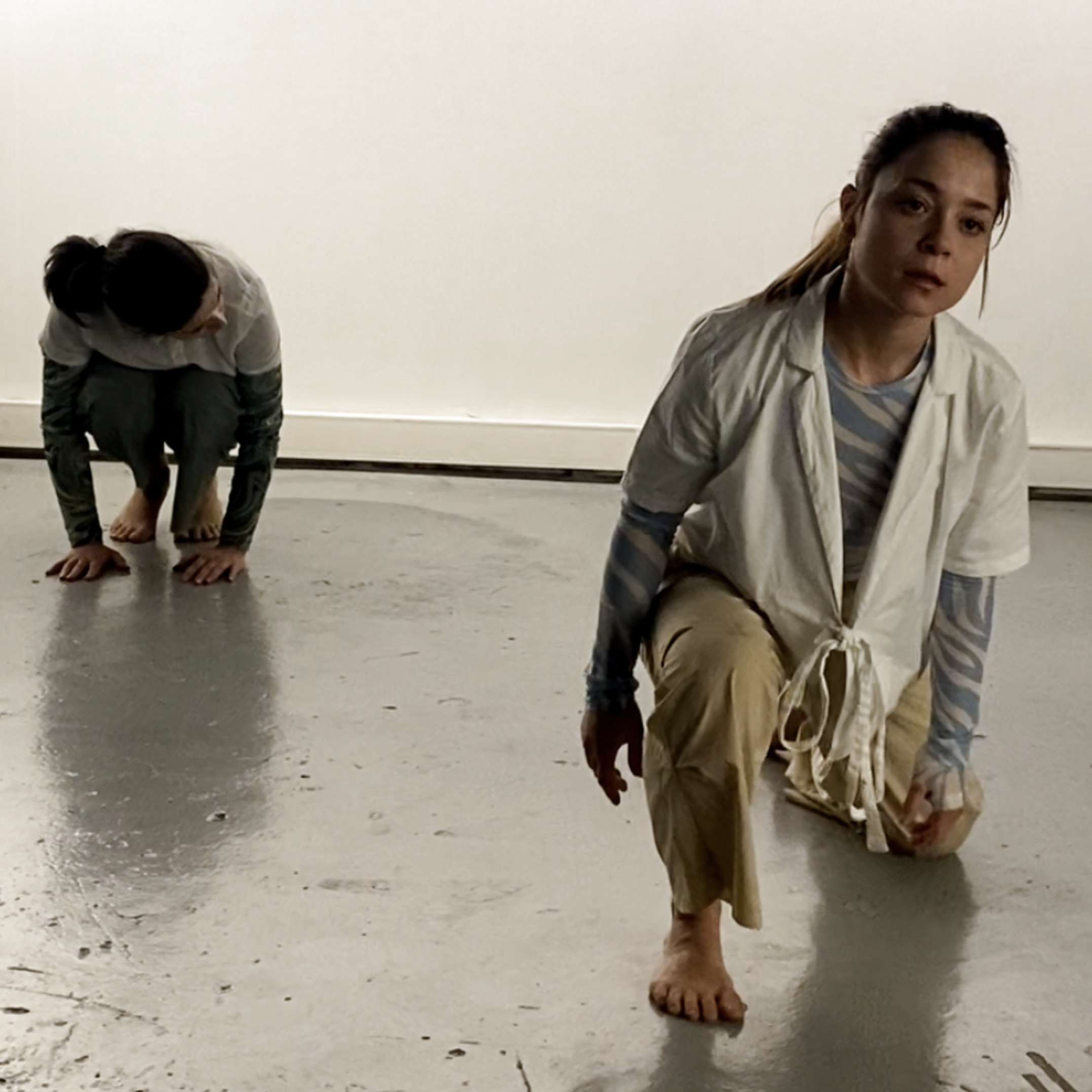 Two women in casual clothing are on the floor in a dance studio, one in the foreground and the other in the background, both in a crawling or crouching position.