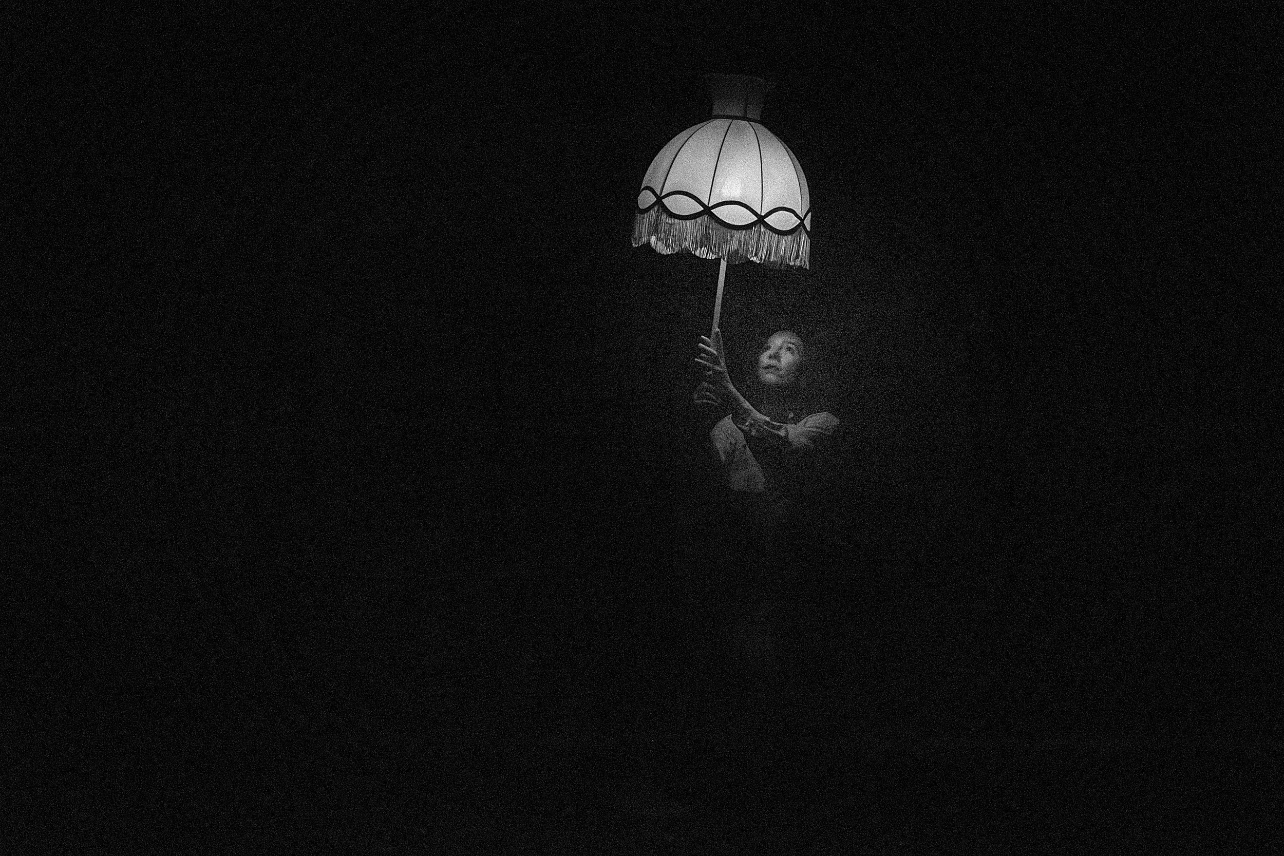 A black and white photo of a puppet or puppet-like doll holding a paper lampshade with fringe, illuminated in a dark setting.