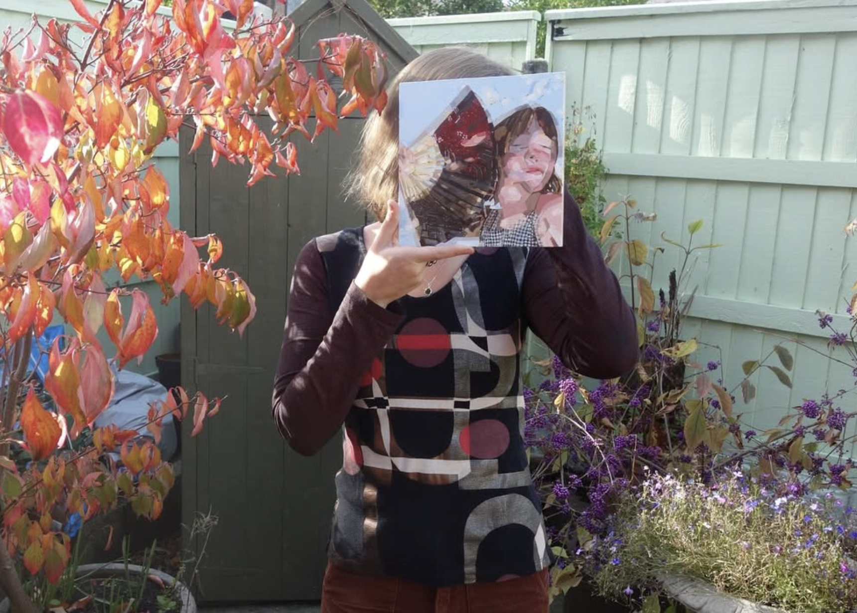Person holding a reflective mirror up to their face, standing outdoors in a garden with plants and flowers, and a wooden fence in the background.