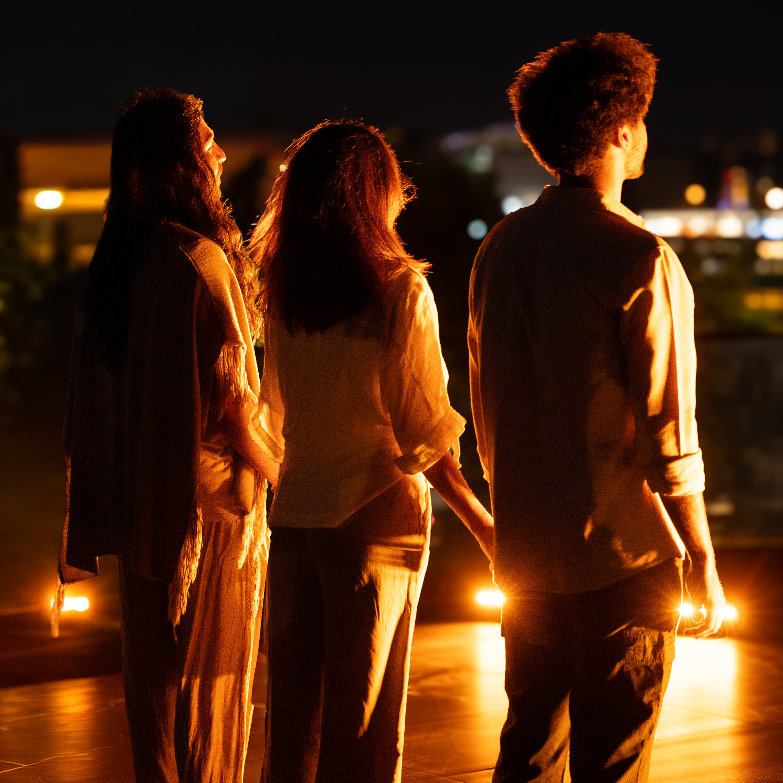 Three people standing on a balcony at night, illuminated by warm orange lights with a cityscape in the background.