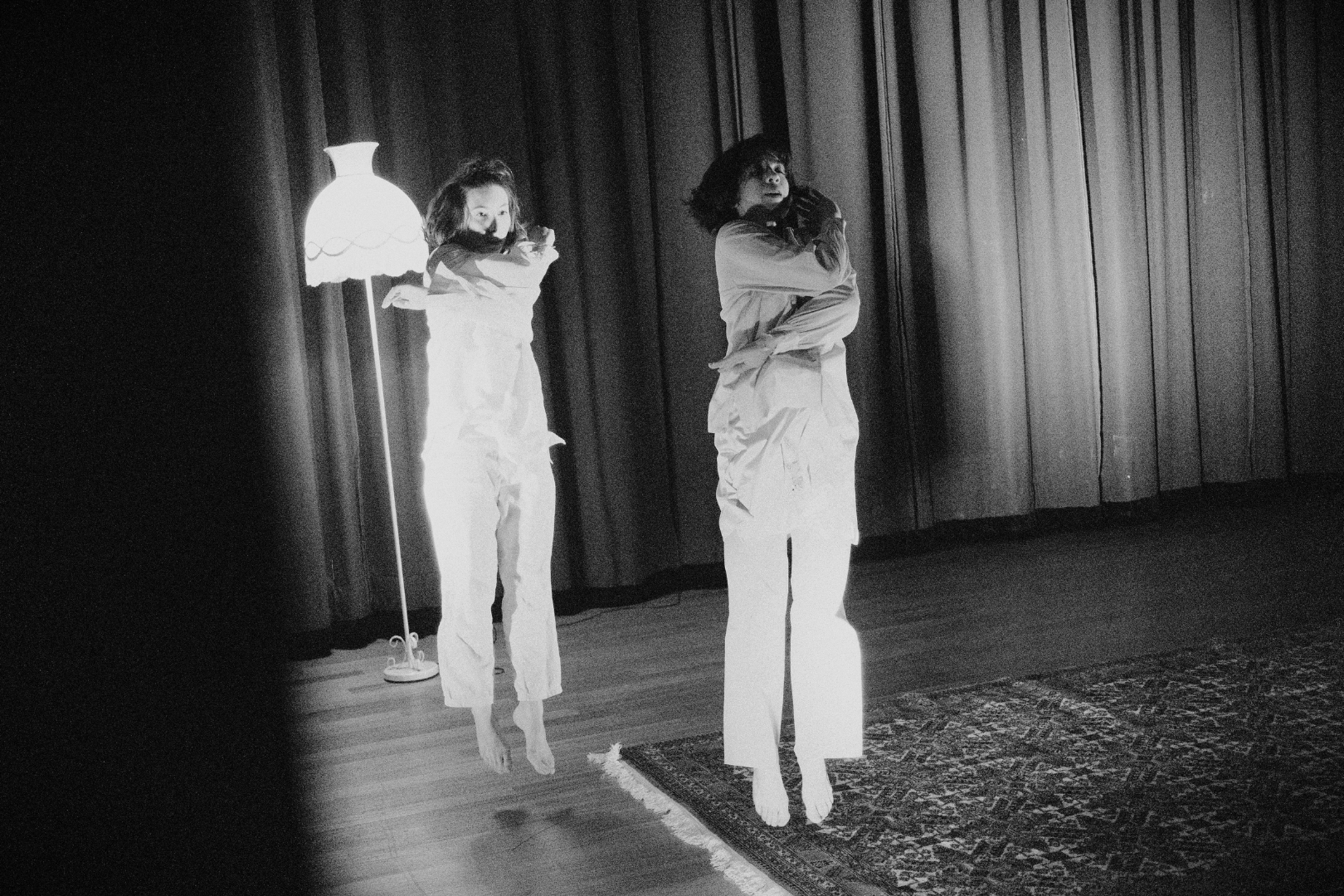 Two women stand on a stage with curtains in the background, wearing white outfits. One woman is standing on the floor, while the other appears to be levitating or jumping, and there is a floor lamp illuminates from behind.