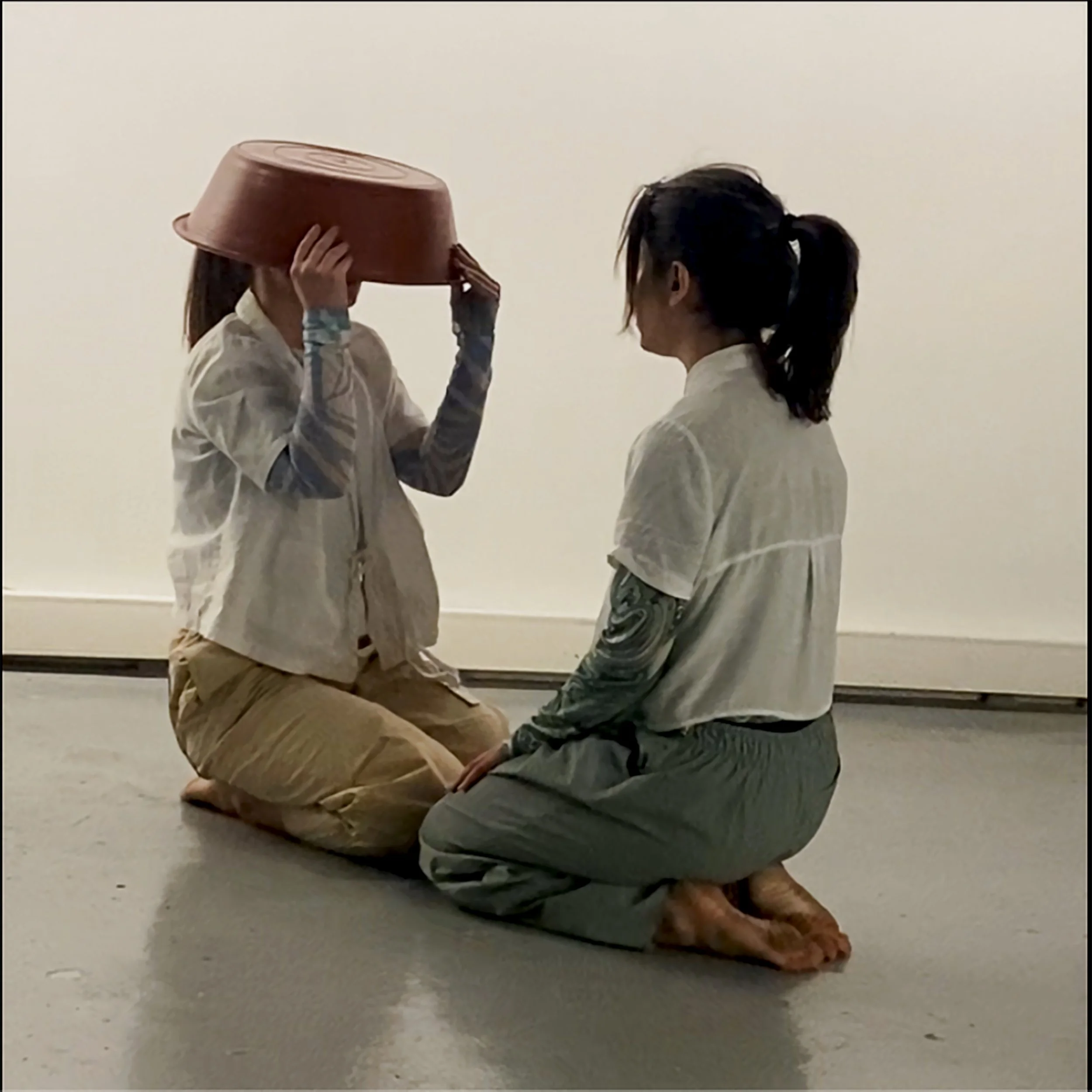 Two young women kneeling on the floor with one wearing a plastic bowl on her head and the other smiling at her, in front of a plain white wall.