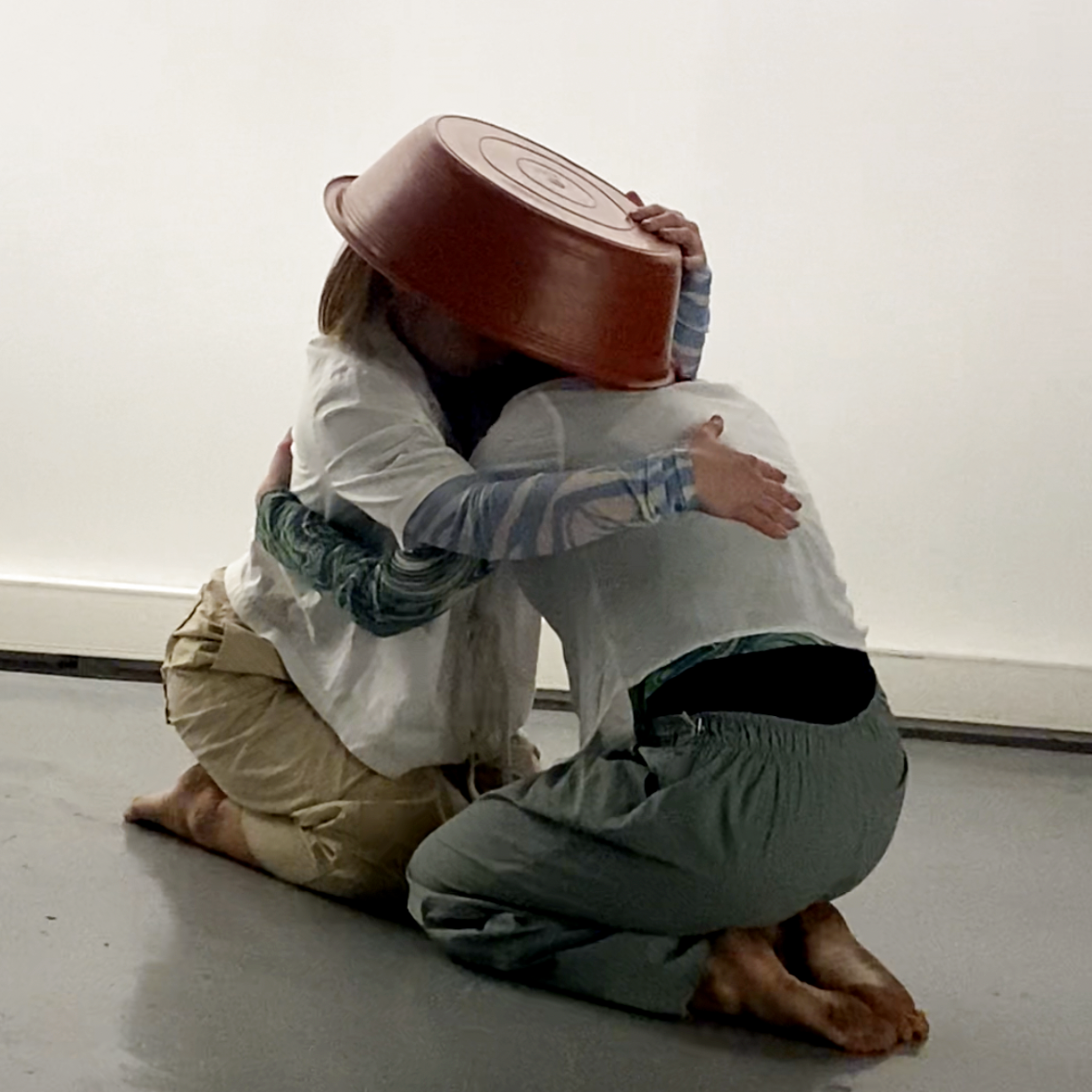 Two people are sitting on the floor and hugging each other. One person is wearing a white shirt and beige pants, with a large orange plastic basin on their head. The other person is wearing gray pants and a gray shirt with long sleeves. They are in a plain, white-walled room with a gray floor.