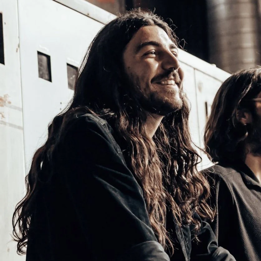 A man with long curly hair, beard, and mustache, smiling with eyes closed, wearing a black jacket, sitting next to a woman with dark hair and glasses, in an indoor setting with a white wall and industrial pipes in the background.