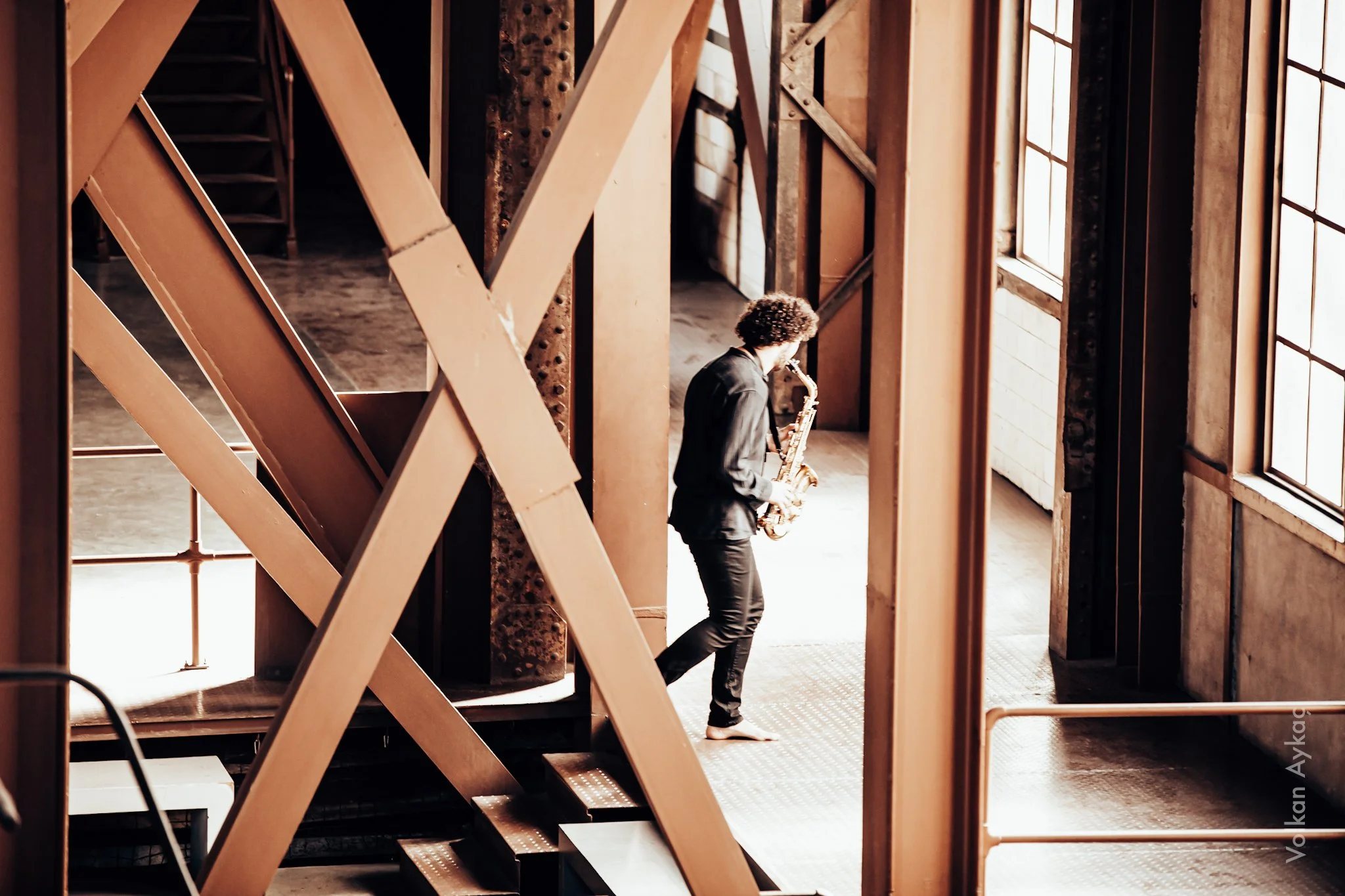 A person with curly hair, dressed in black, barefoot, playing a saxophone in an industrial-style room with large windows and exposed wooden beams.
