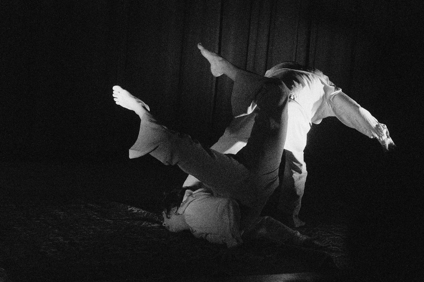 A black and white photo of two people practicing Brazilian Jiu-Jitsu on a dark mat. One person is on top while the other is on their back, both are wearing gis.