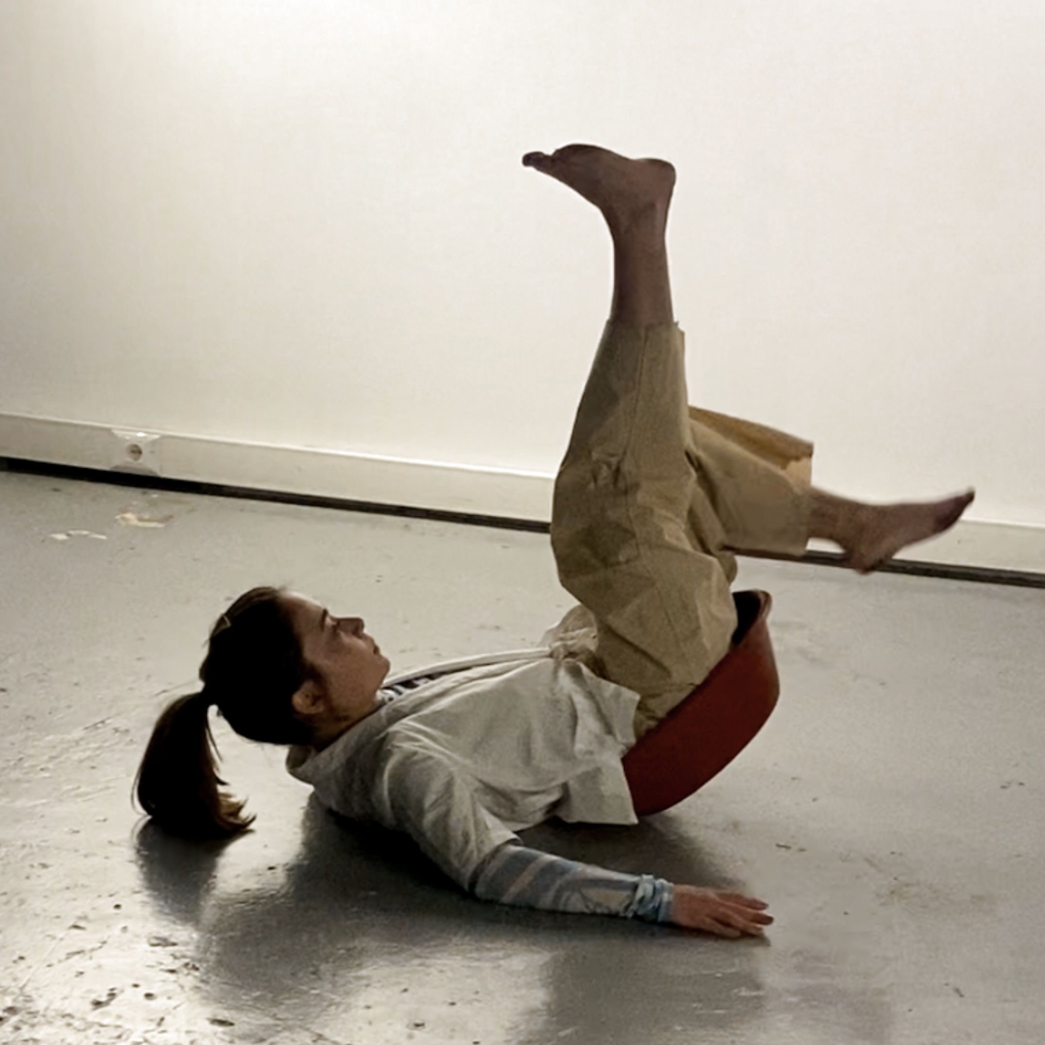 A person lying on their side on the floor with legs raised and crossed, wearing beige pants and a white shirt with tattoos on the arm, in a room with a plain white wall.