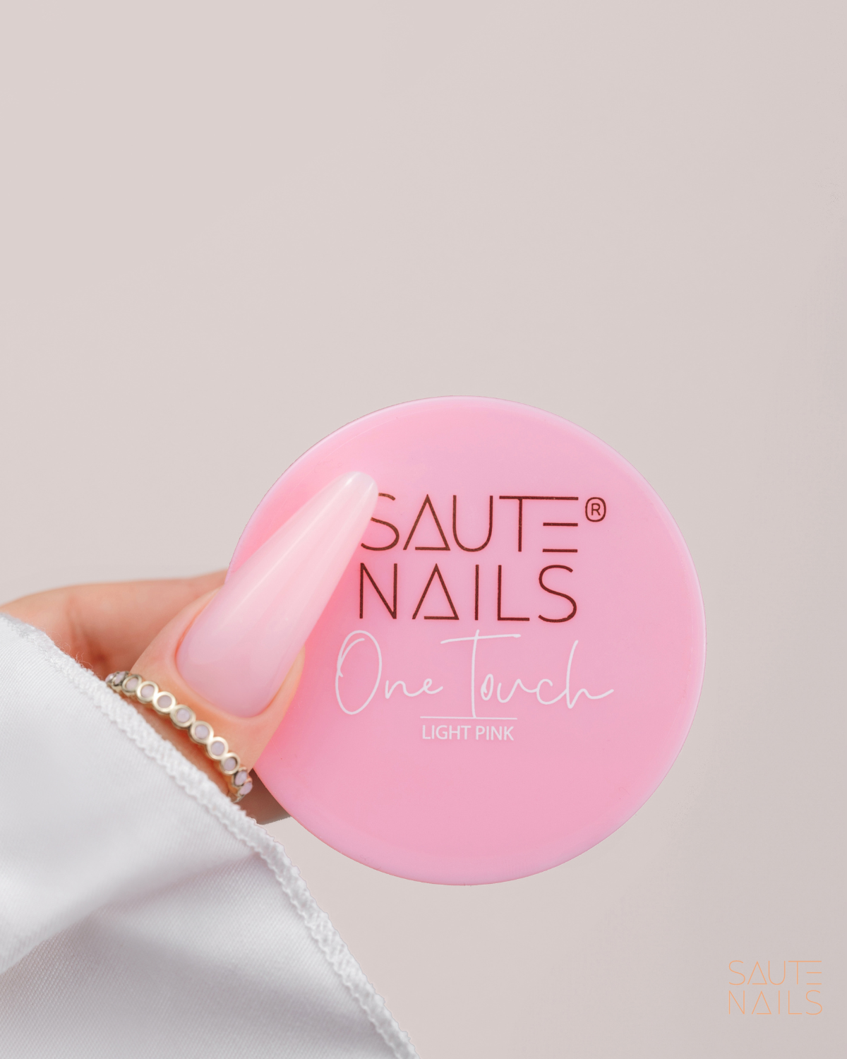 Mano con unghie finte di colore rosa chiaro che tiene una confezione rosa di smalto per unghie con etichetta 'Saute Nails One Touch Light Pink'.