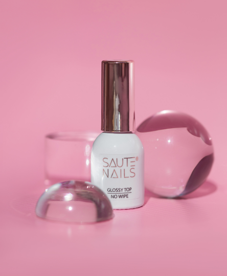 Bottiglia di smalto per unghie bianco con cappuccio argentato, posizionata su una superficie rosa con alcuni oggetti di vetro trasparenti e rosa intorno.