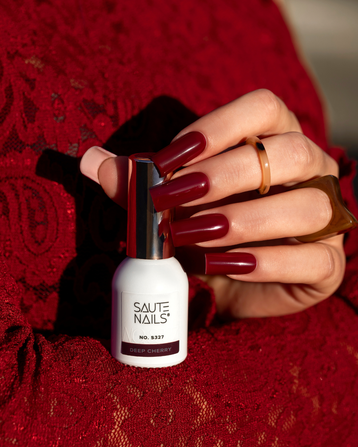 Mano con unghie dipinte di rosso scuro che tiene una bottiglia di smalto per unghie di colore bianco con etichetta "Saute Nails Deep Cherry"