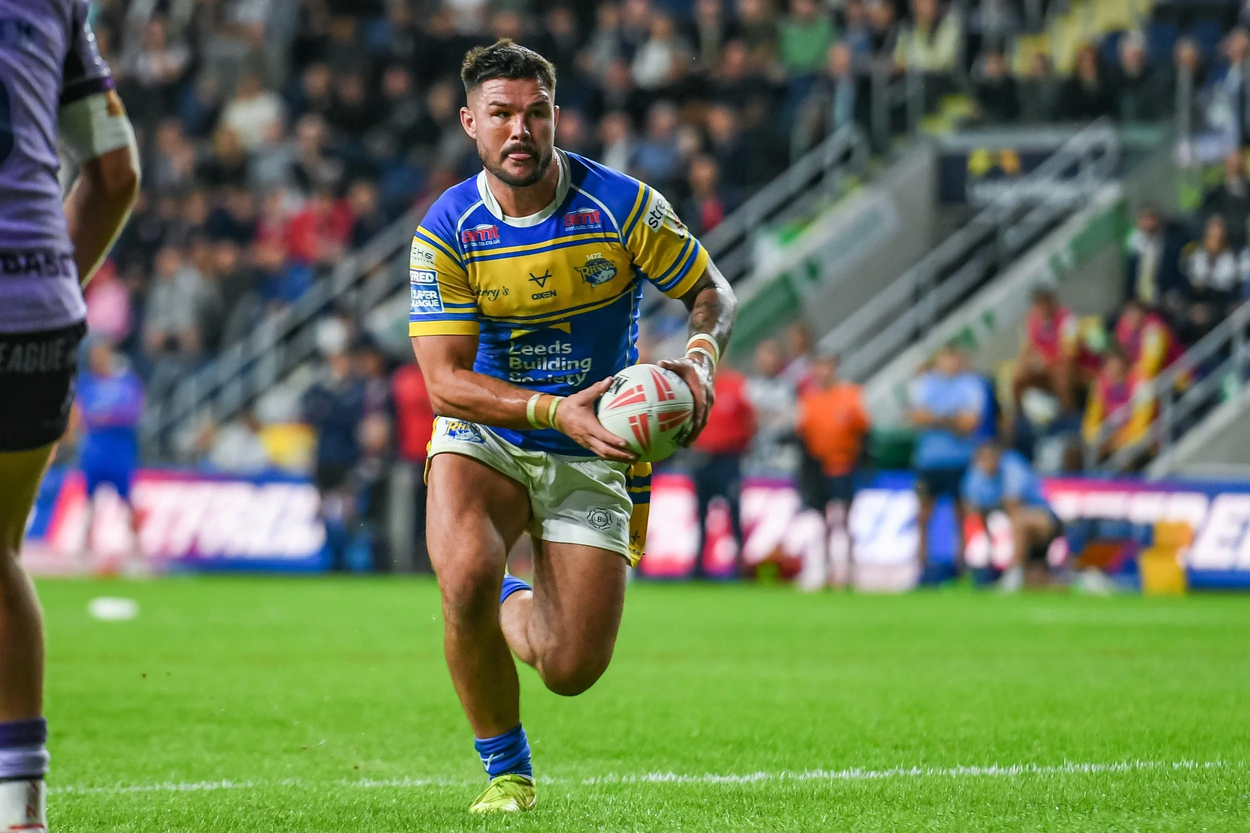 A rugby player in a yellow and blue uniform running on a field, holding a rugby ball, with spectators in the background.