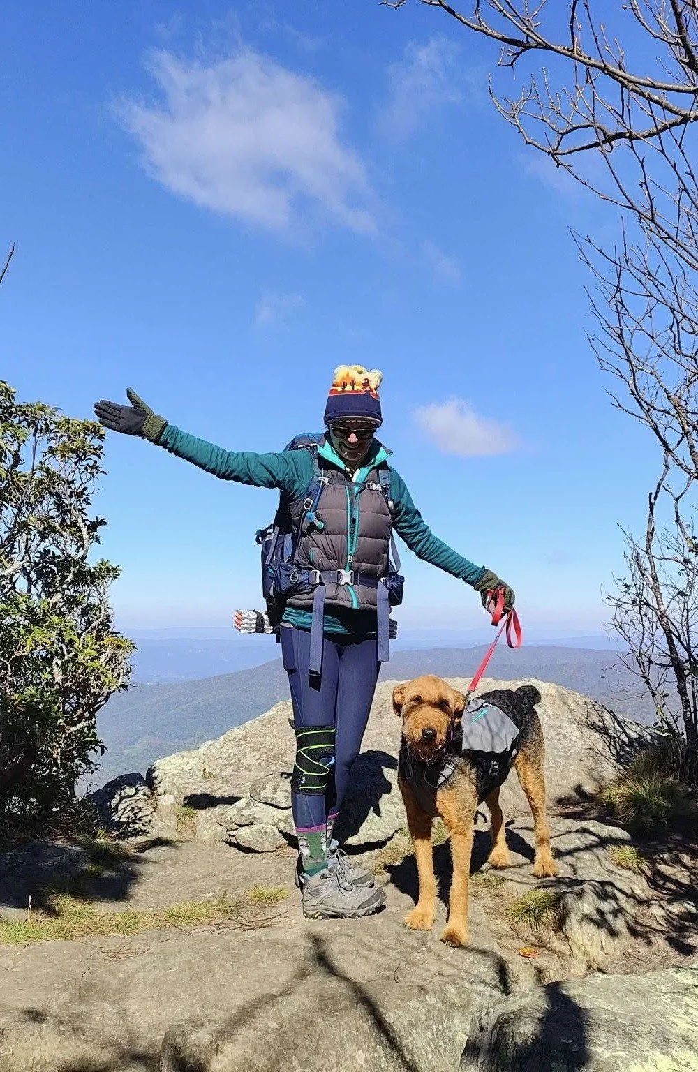A hiker and her dog on a mountain summit, with blue sky and scattered clouds in the background.