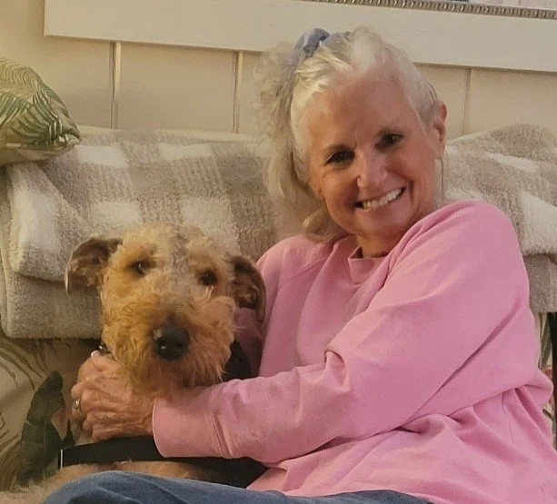 A smiling woman with white hair, wearing a pink sweatshirt, sitting on the floor and hugging a tan and black dog with a scruffy coat indoors.