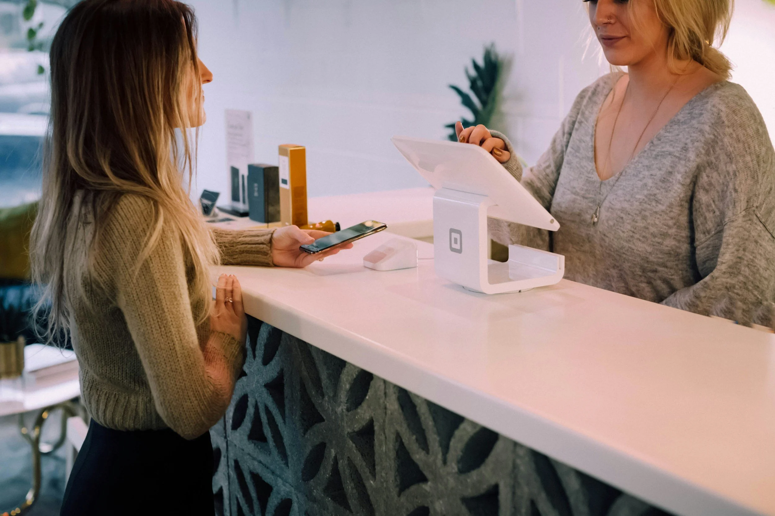 A customer at a counter making a payment using a smartphone while a cashier operates a point-of-sale system in a modern, well-lit space.