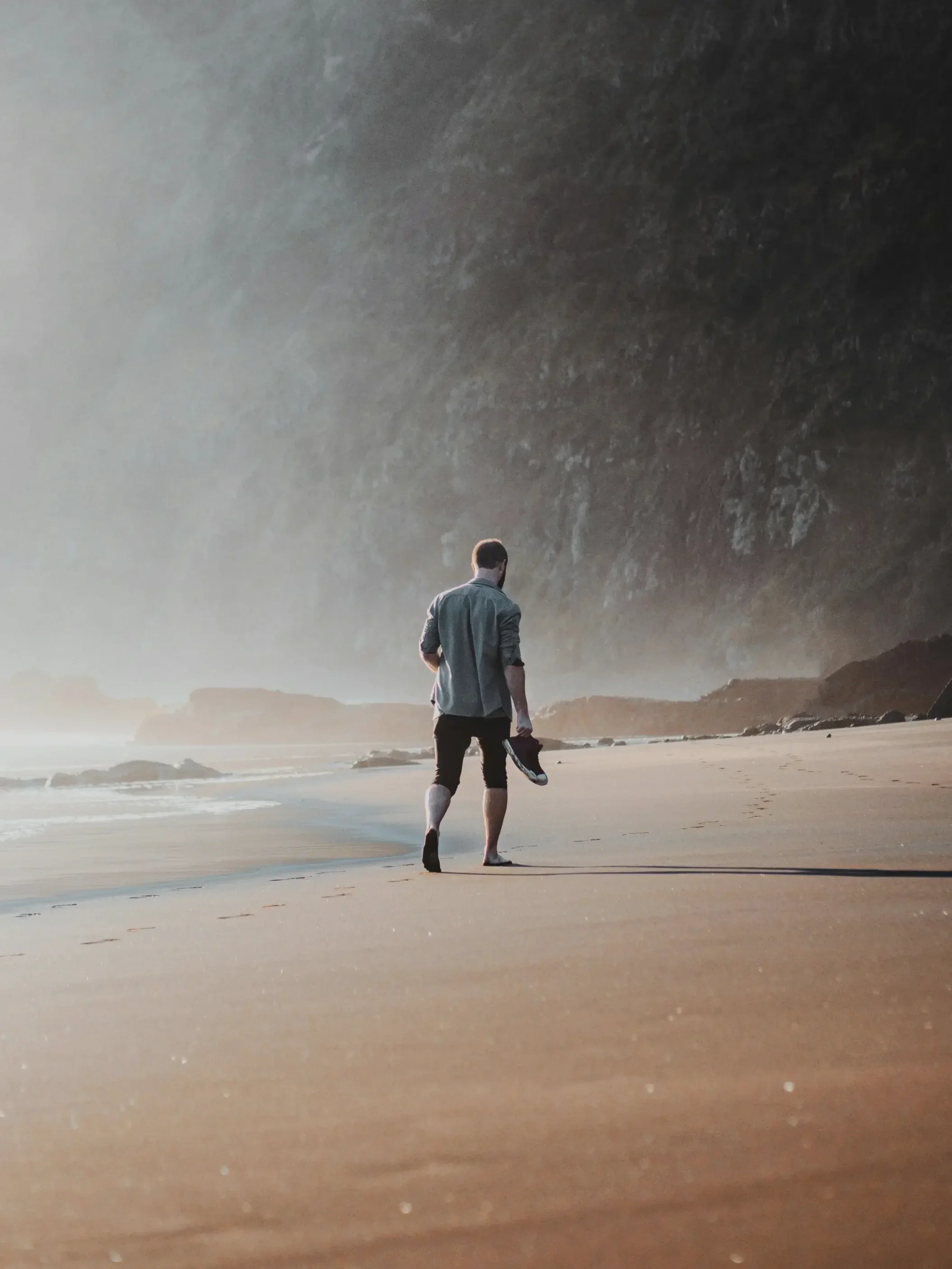 A man walking on a sandy beach with the ocean and cliffs in the background, carrying shoes in his hand.
