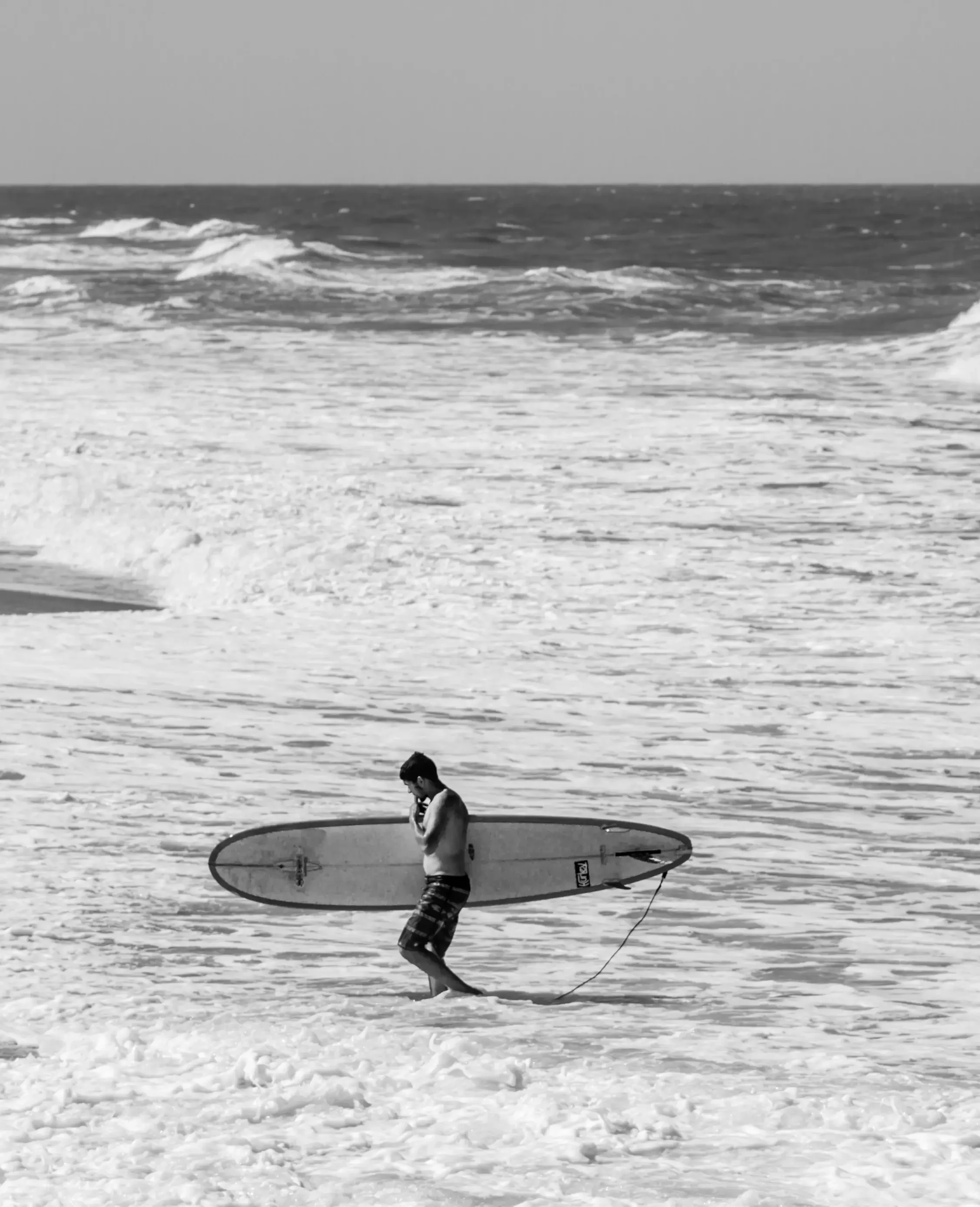 A young man in swim shorts carrying a surfboard walks in shallow water at the beach.