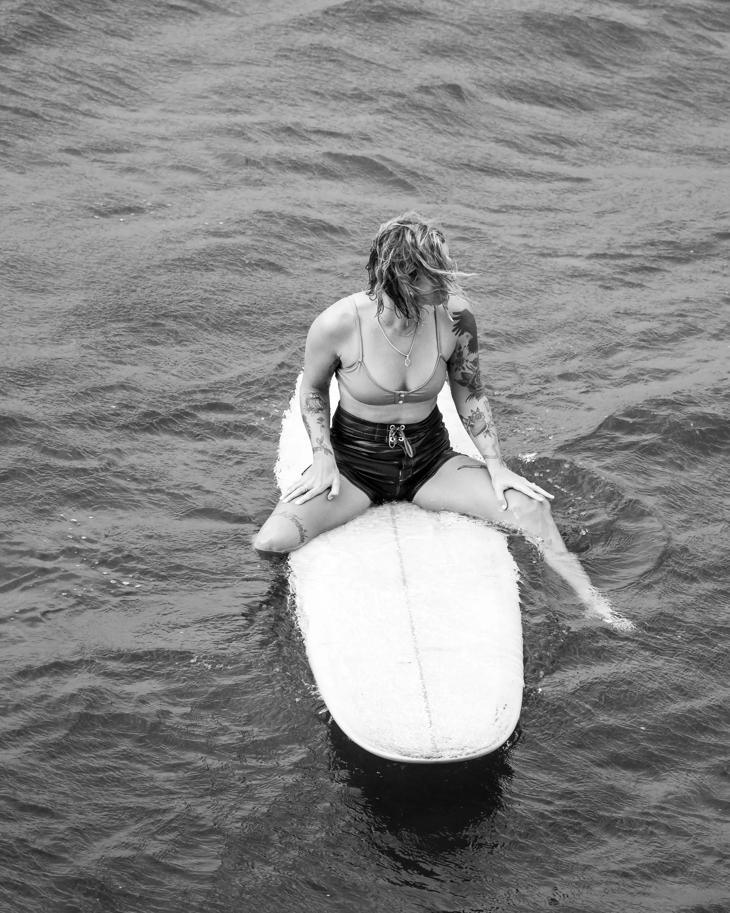 A woman with tattoos and wet hair sitting on a surfboard in the water.