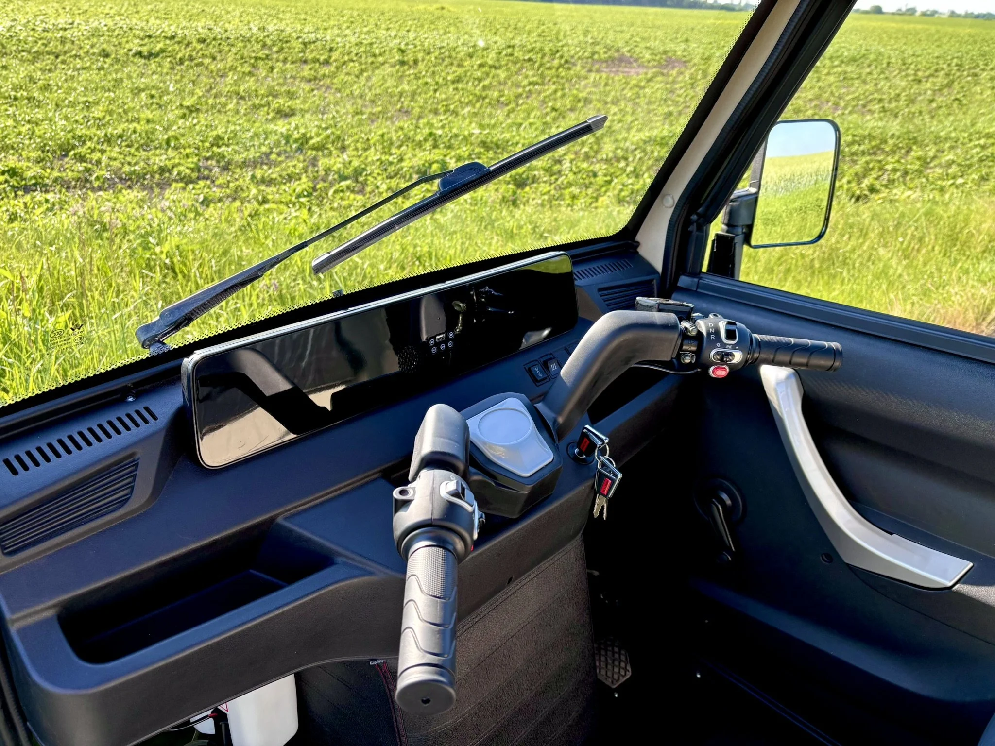 Indre kabine i en grøn luxcap el-kabinescooter med styr og instrumentbræt, omgivet af et åbent landskab med græs og marker.