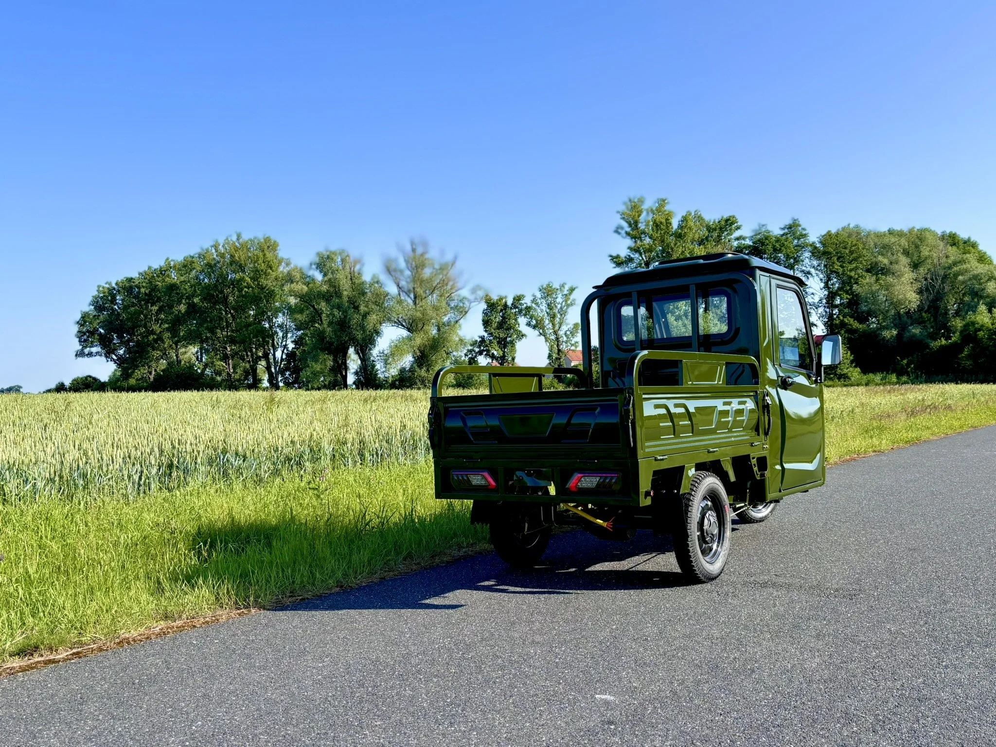 En grøn luxcap el-kabinescooter parkeret på en landevej ved en mark med træer i baggrunden under en klar blå himmel.